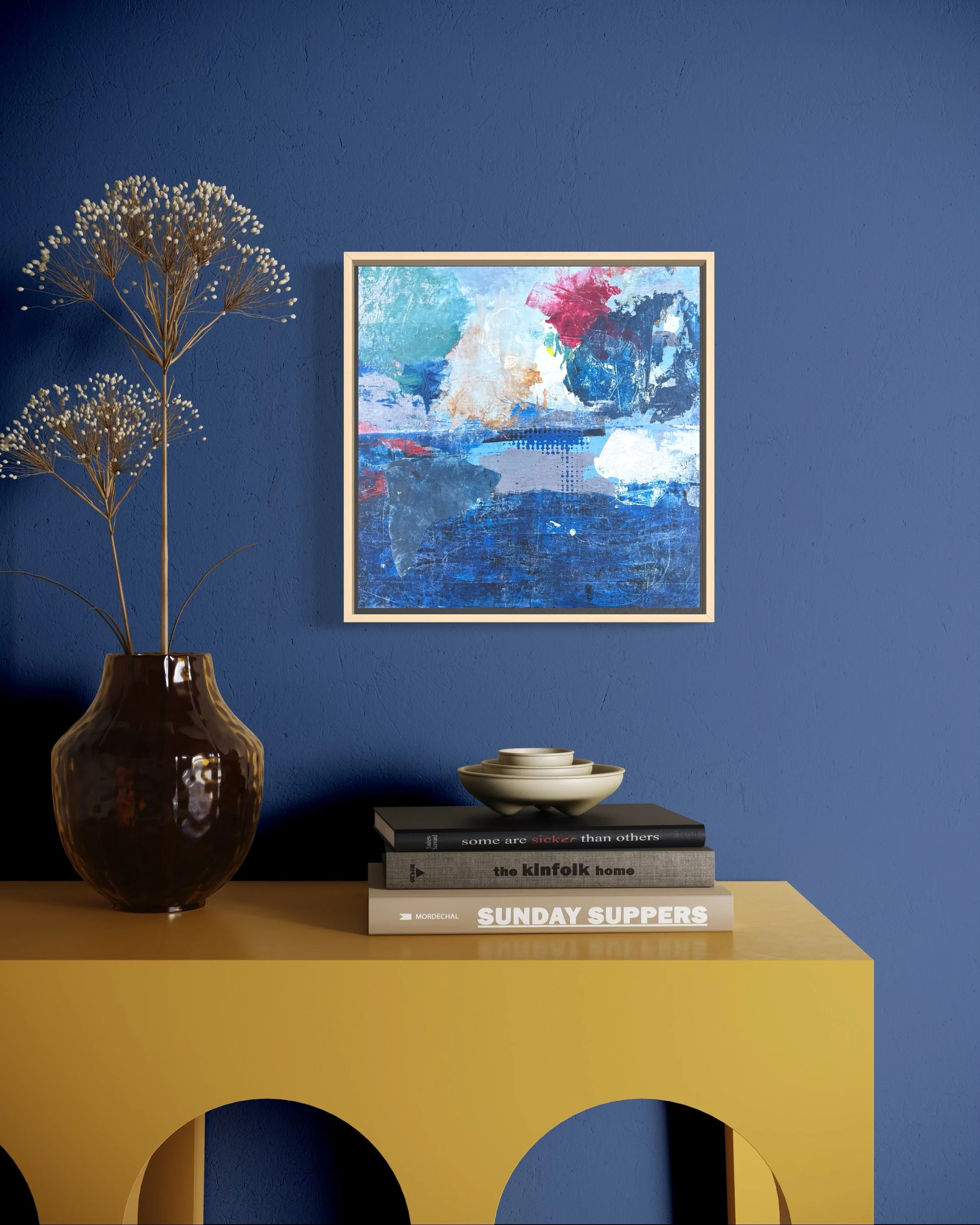 Interior decor with a yellow table, a dark brown vase with dried flowers, and a stack of books topped with a beige bowl, against a blue wall with abstract artwork.