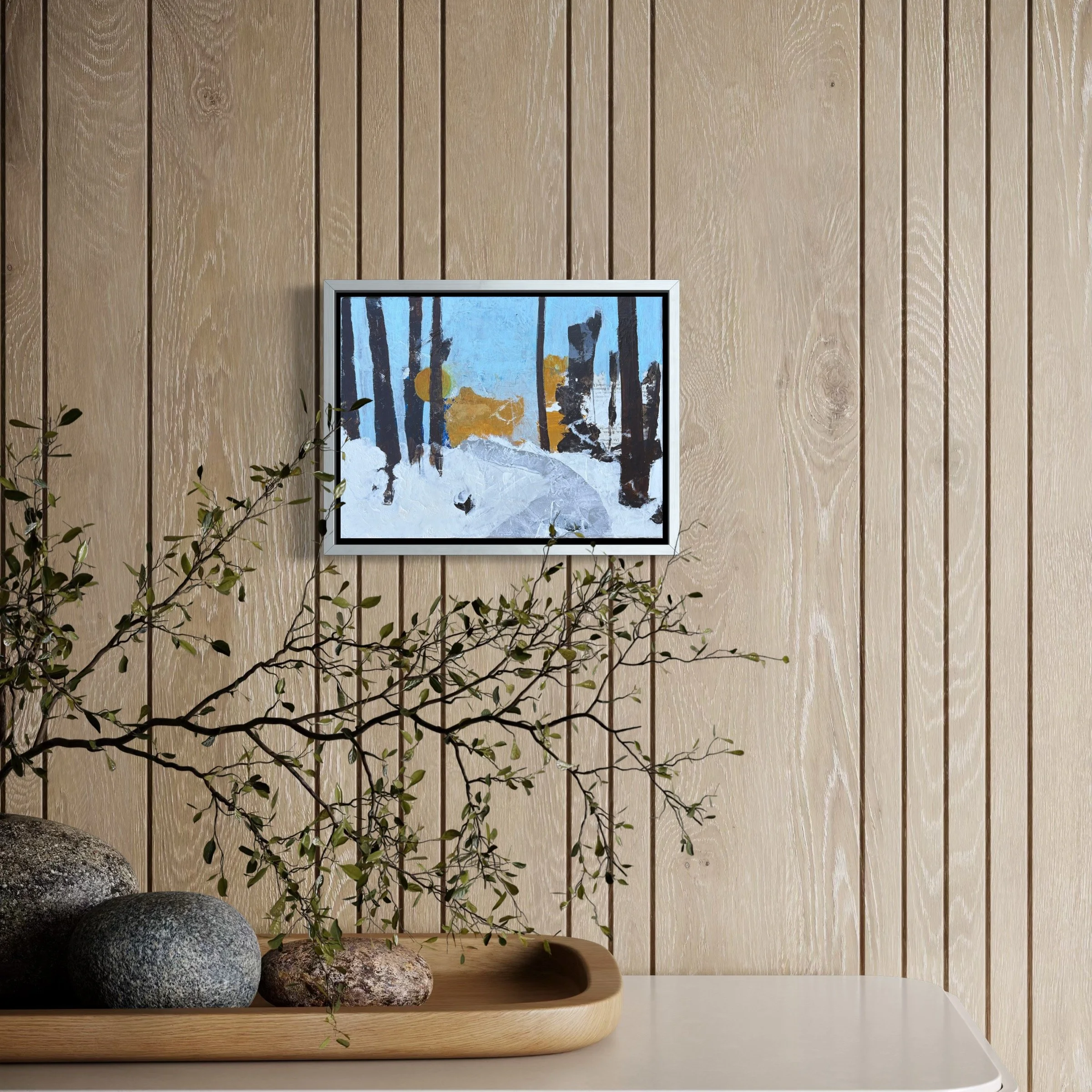 A framed painting hanging on a wooden paneled wall, featuring a winter scene with snow, trees, and a blue sky. Below, a wooden tray with four rocks and a small leafy branch. A white table surface is partially visible.