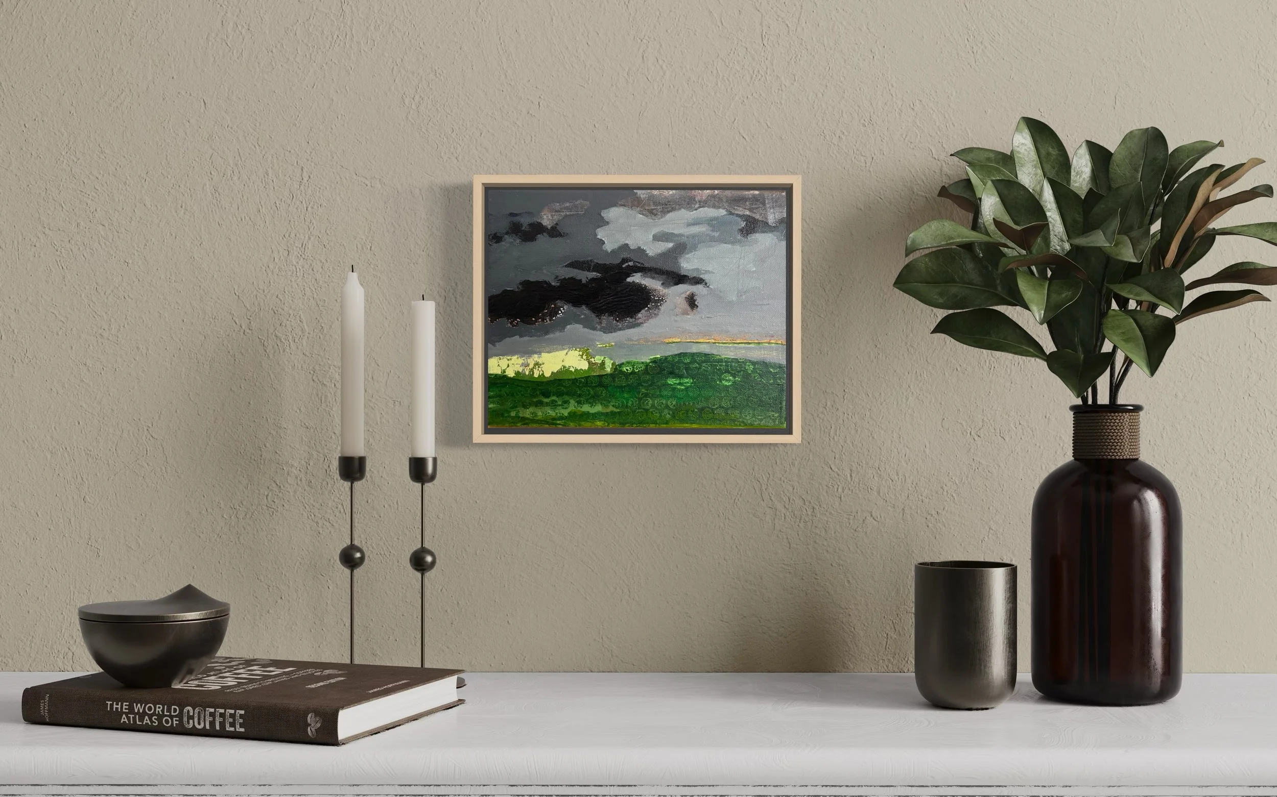 Decorative table with a black bowl, a large coffee table book, two white candles in black holders, a small black cup, and a large dark brown vase with green leaves, against beige textured wall with a framed landscape painting.
