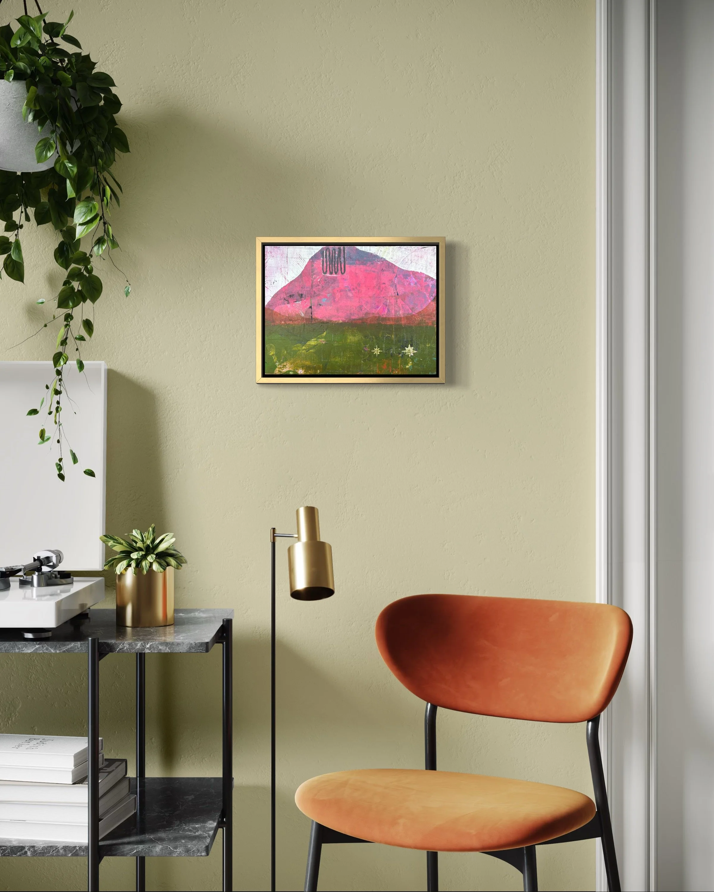 A cozy room corner with a green wall, a framed abstract pink and green painting, an orange cushioned chair, a small black metal table with books and a plant, and a tall brass floor lamp.
