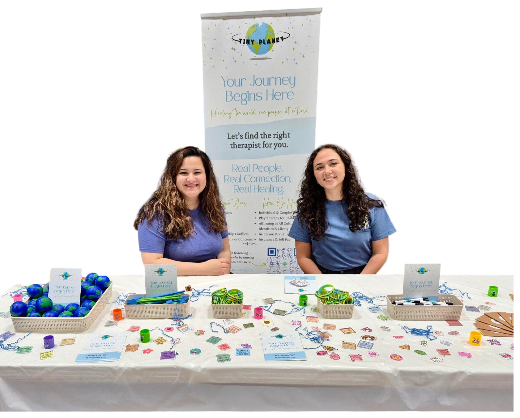 Two women smiling behind a table with promotional items and signs, set up for a health or wellness event, with a banner behind them displaying the text 'Your Journey Begins Here' and information about finding a therapist.