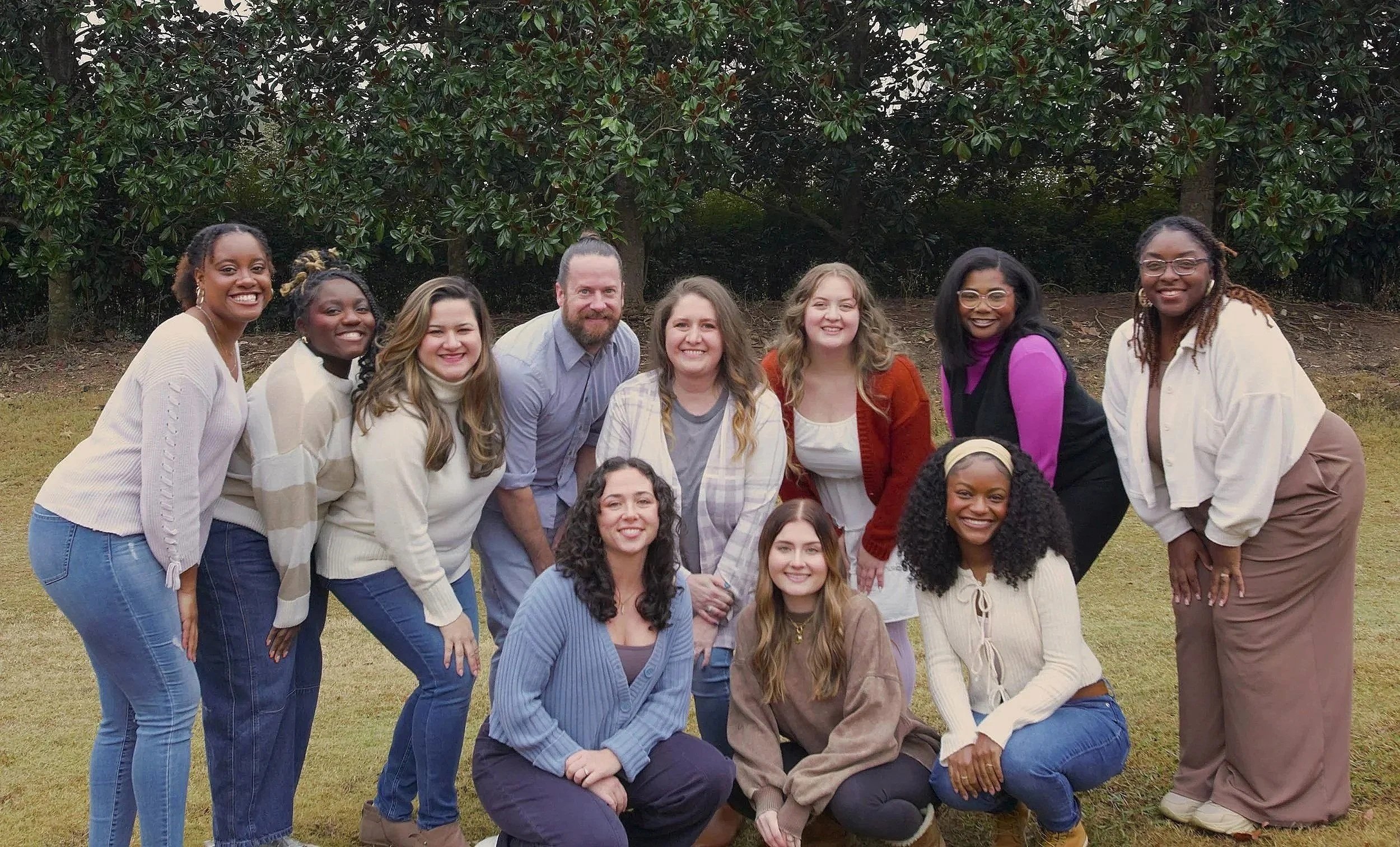 A group of 12 people, including men and women of diverse ethnicities, smiling and posing outdoors on a grassy area with trees in the background.