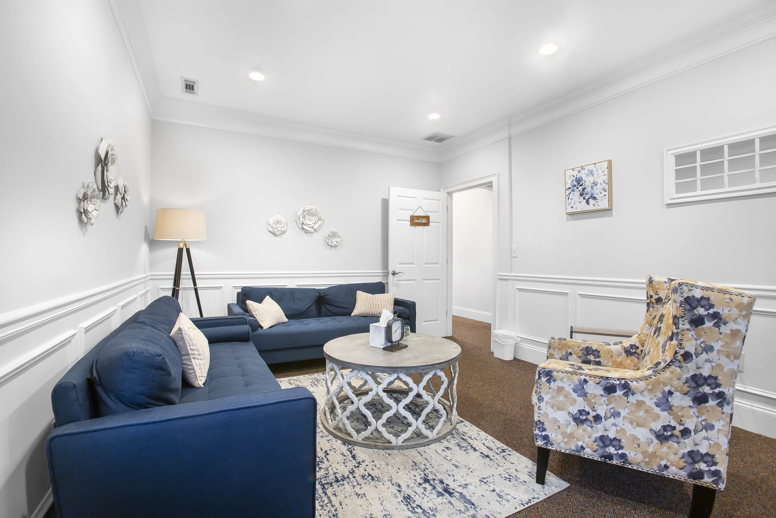Living room with white walls, two blue sofas with pillows, a floral patterned armchair, a round coffee table, a floor lamp, wall art, and a small window.