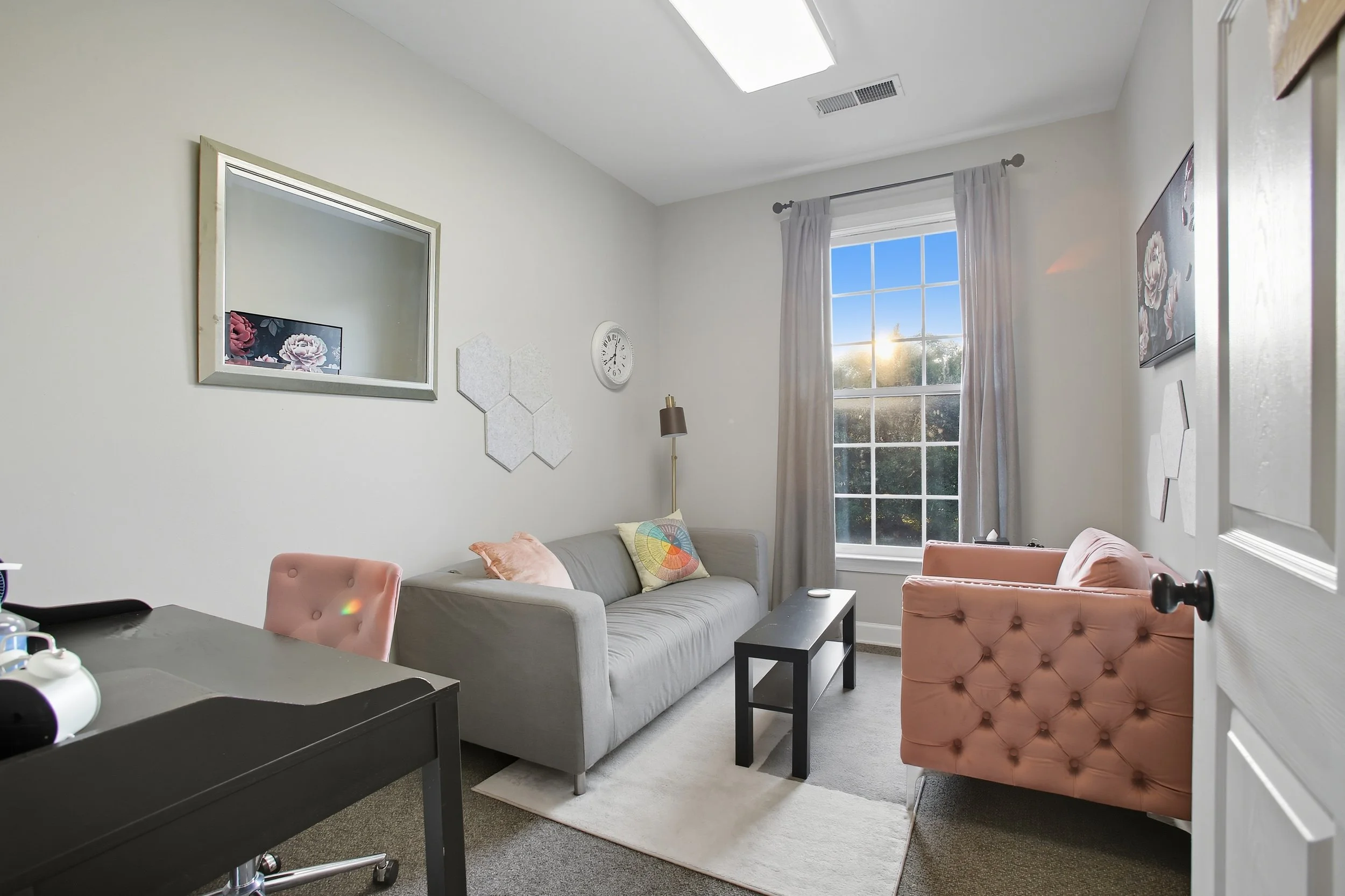A bright living room with a gray sofa, pink armchair, and black coffee table, illuminated by sunlight through a large window with white curtains, and decorated with wall art and a clock.