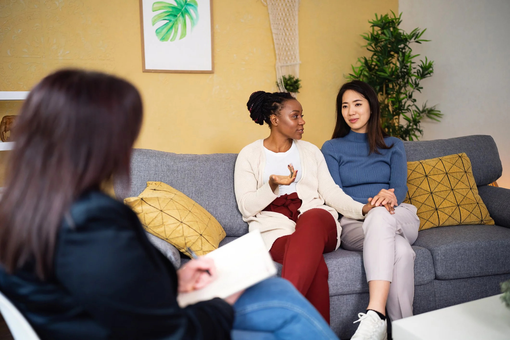 Three women having a conversation in a living room with yellow walls, a blue couch, yellow pillows, and potted plants in the background.