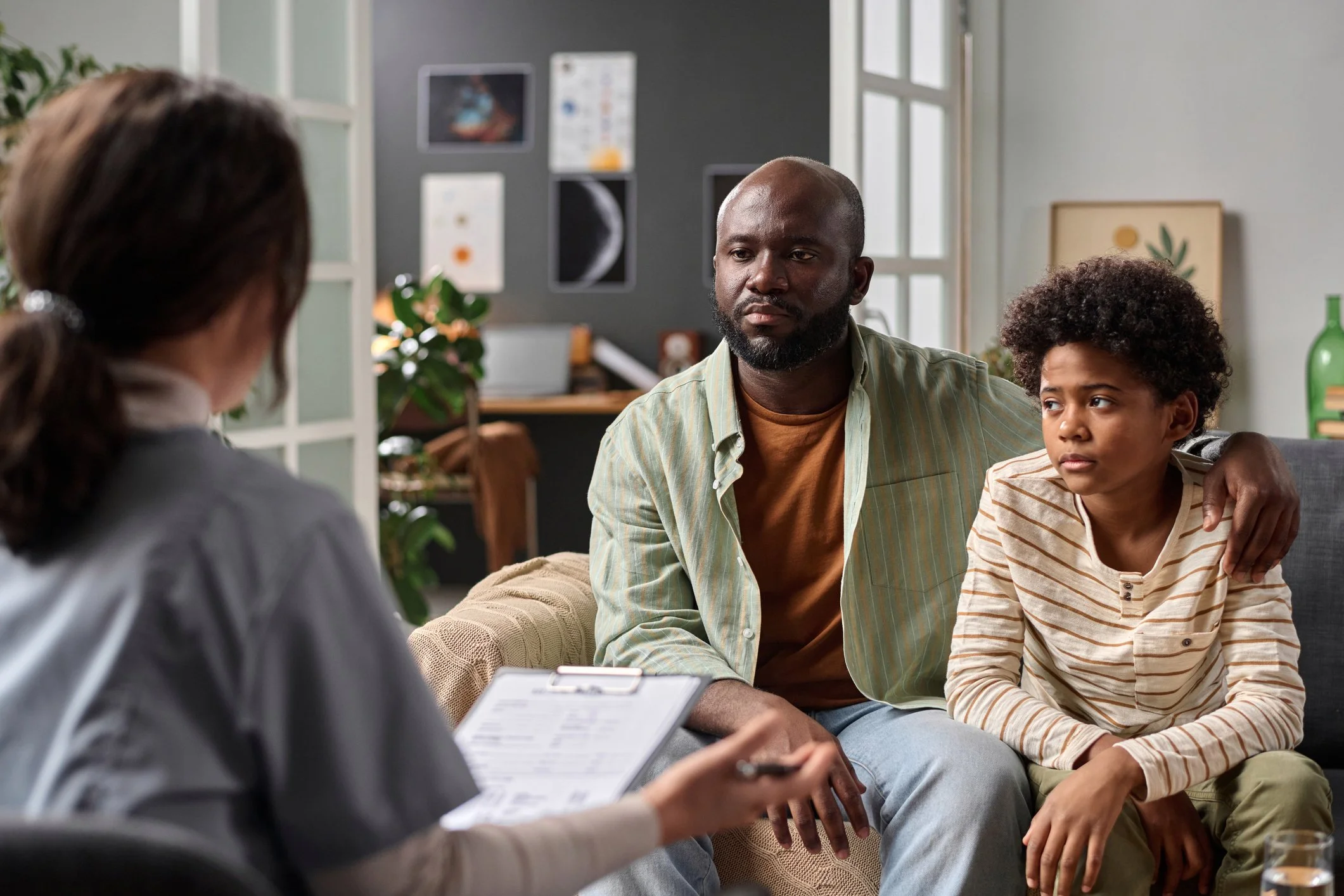 A man and a boy sit on a couch during a therapy session with a therapist in a living room.
