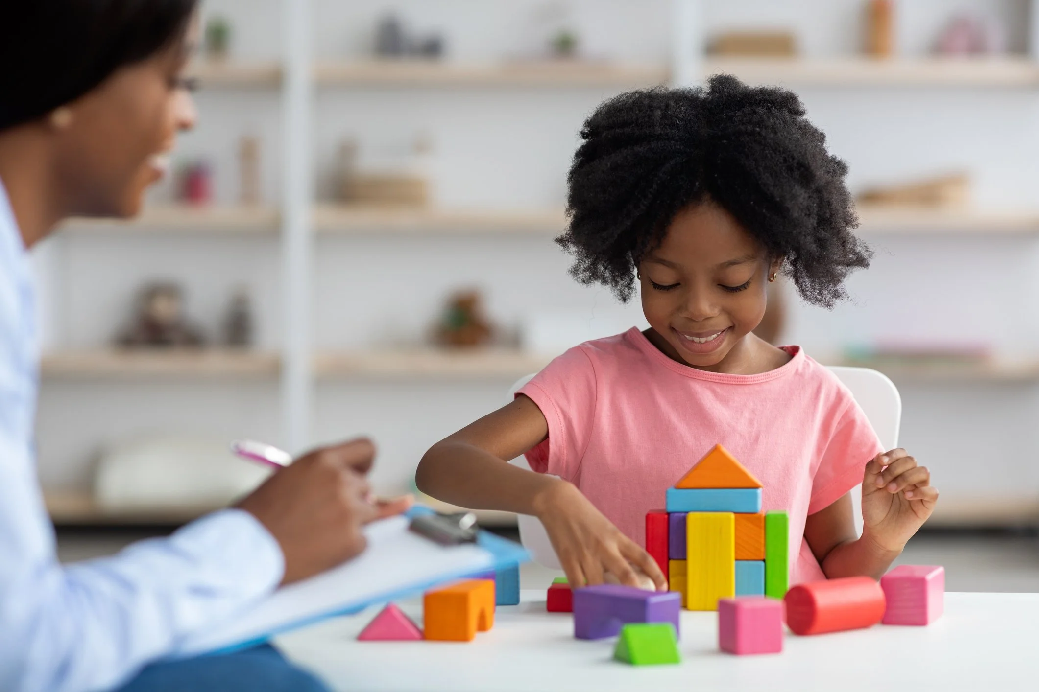 A young girl with curly hair in a pink shirt playing with colorful building blocks at a table, while an adult watches nearby in a bright room.