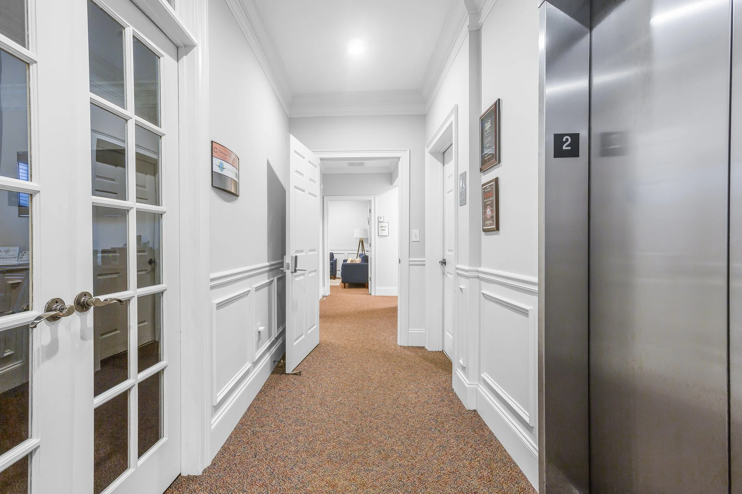 Hallway with open door shows lounge area with sofa and floor lamp at the end. Multiple signs and plaques on white walls, elevator door to the right, carpeted floor, and glass-paned door on the left.