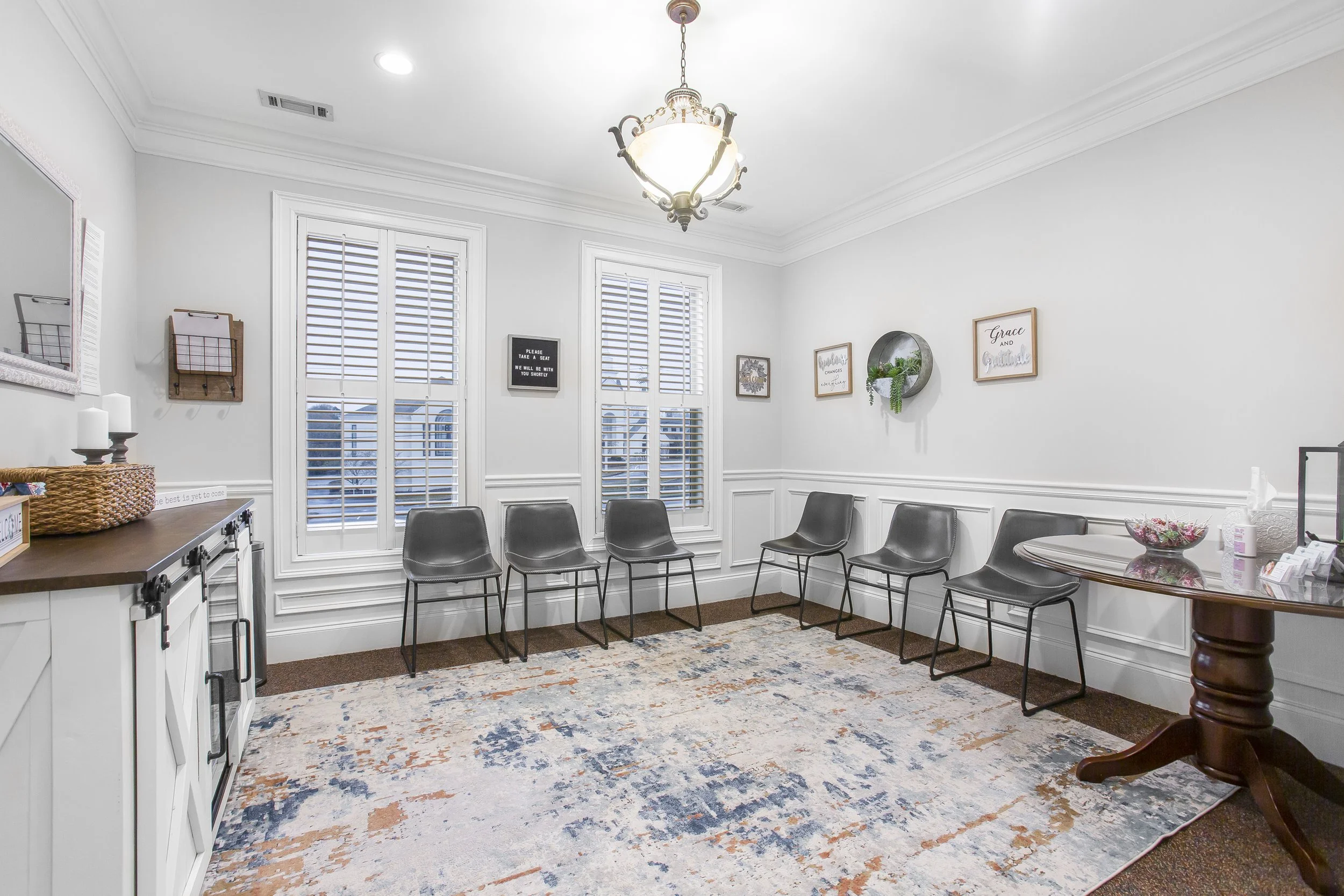 Empty waiting room with six black chairs along the wall, decorative framed signs, a chandelier, white walls, and large windows with shutters.