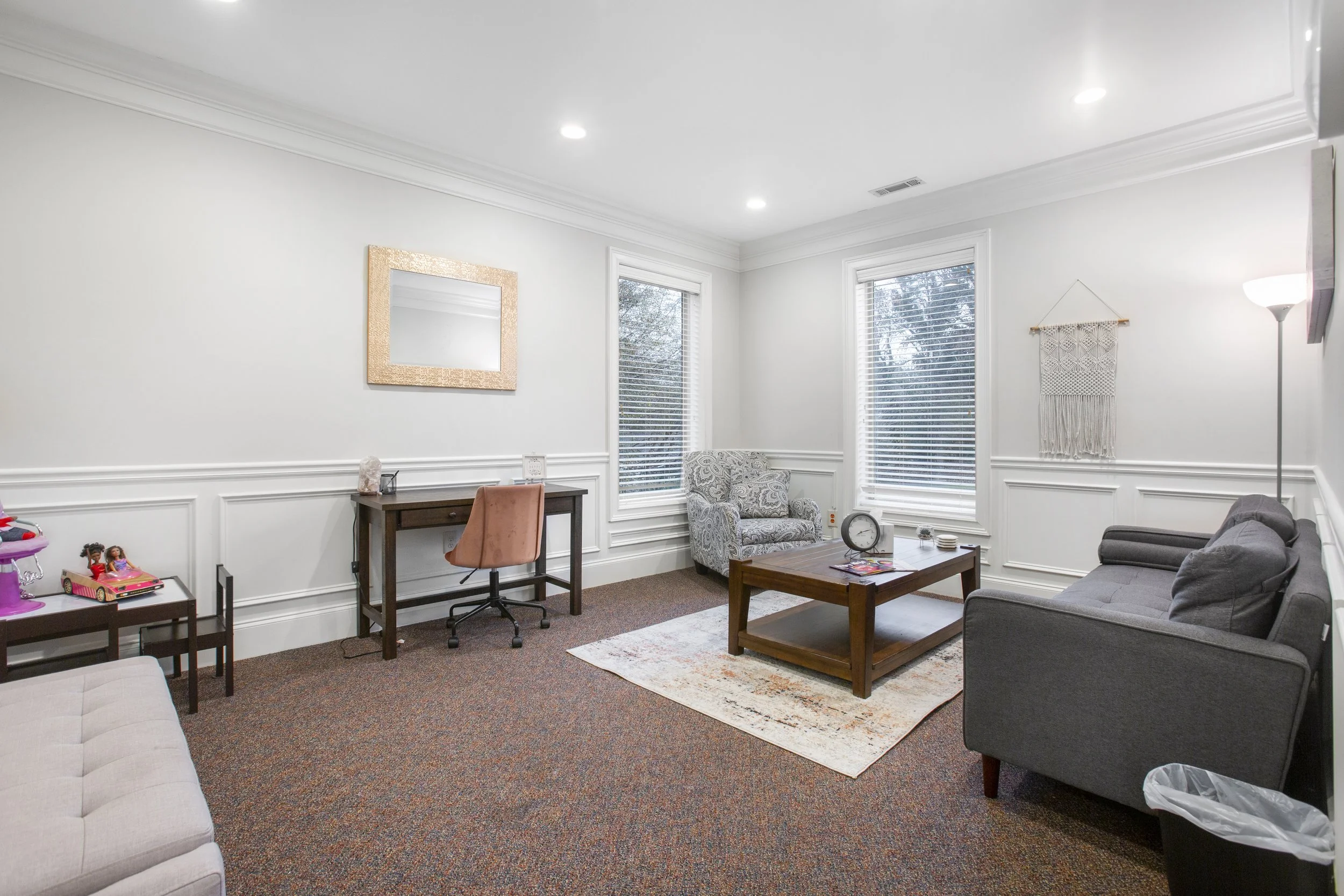 Living room with white walls, three large windows with blinds, a gray sofa, a patterned armchair, a wooden coffee table, a desk with a pink chair, and children’s toys on a small table.