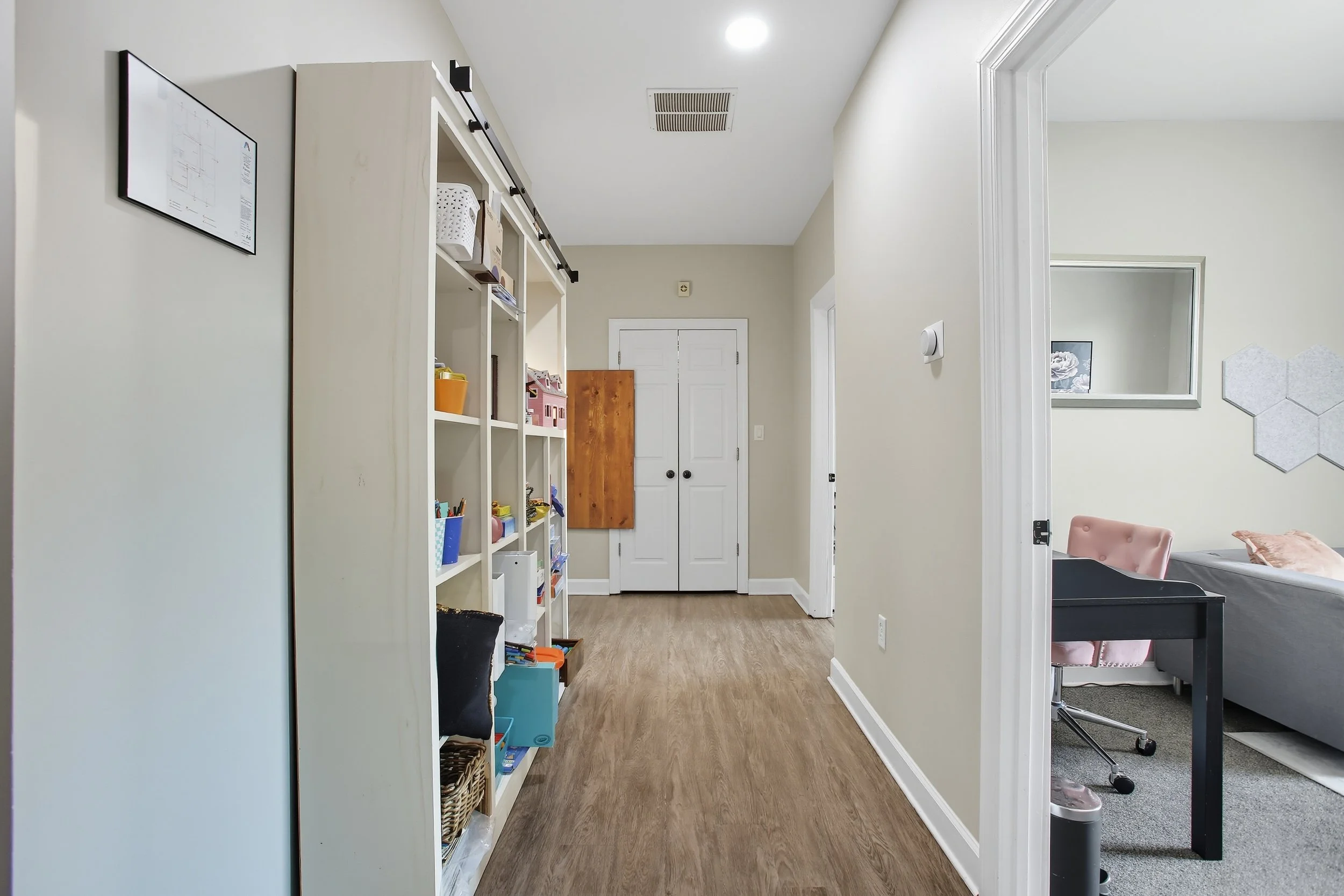 Interior view of a hallway with white walls, a wooden floor, a white bookshelf filled with toys and books, and a closed white door at the end. To the right, an open doorway reveals a room with a desk, pink chair, couch, and wall decorations.