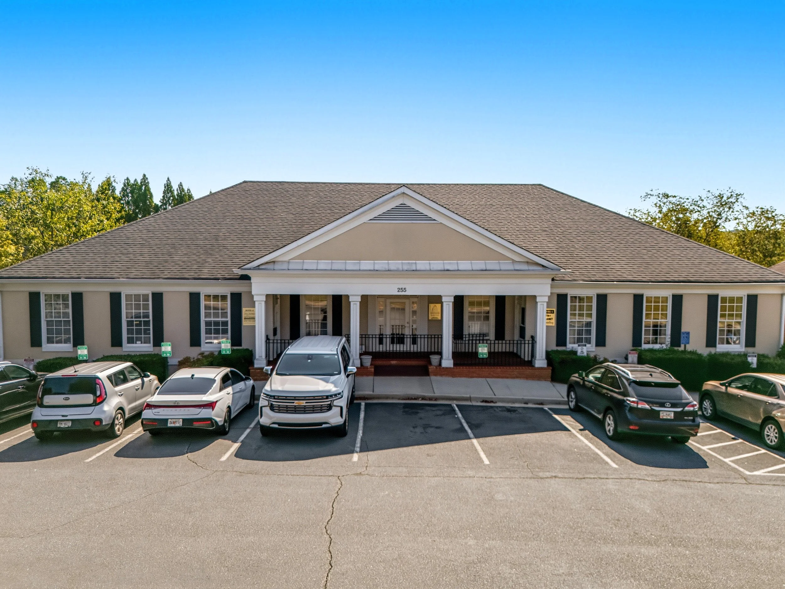 A single-story beige building with white columns and a gabled roof, surrounded by a parking lot with several cars parked in front.