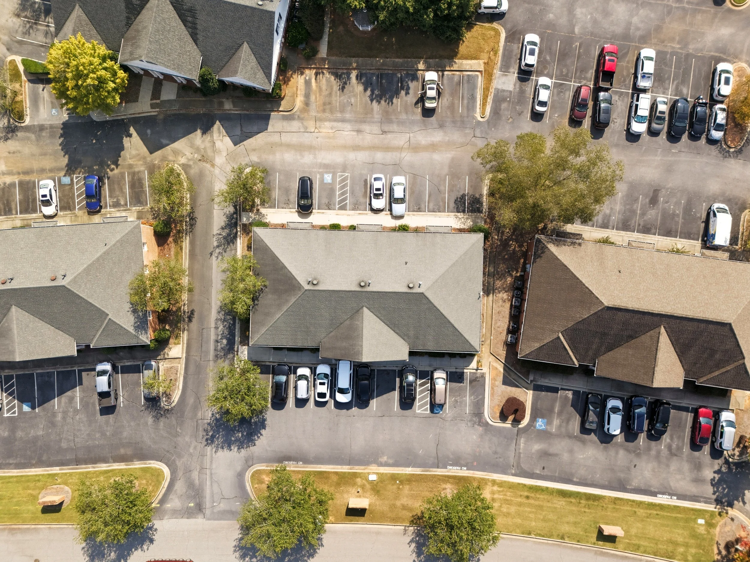 Aerial view of a parking lot adjacent to residential buildings with multiple parked cars and trees.
