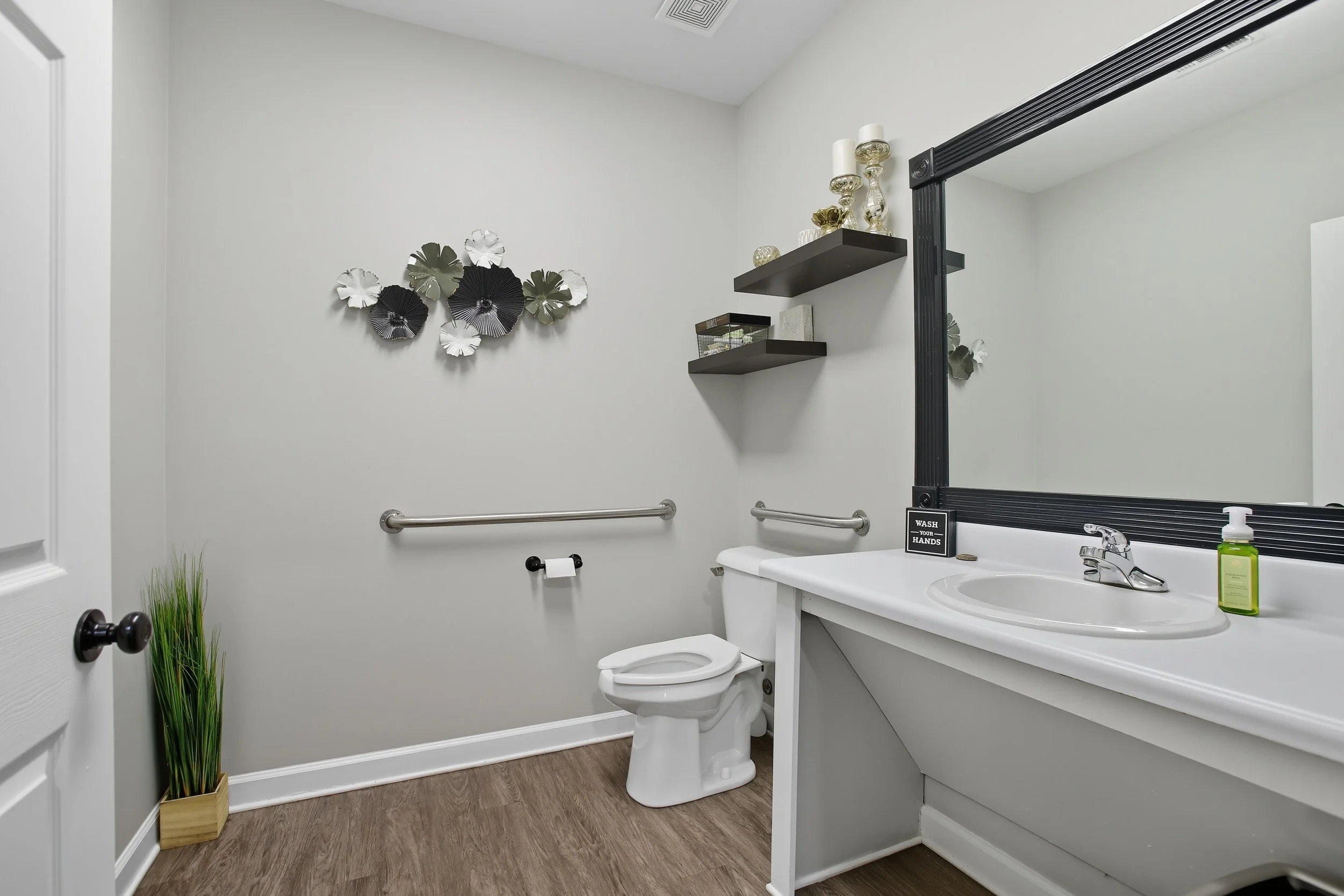 A clean, modern bathroom with a white toilet, a large mirror with a black frame, a white sink, and dark wall-mounted shelves decorated with candles and small decorative items. The wall also has black, white, and gray floral wall art. A small potted p