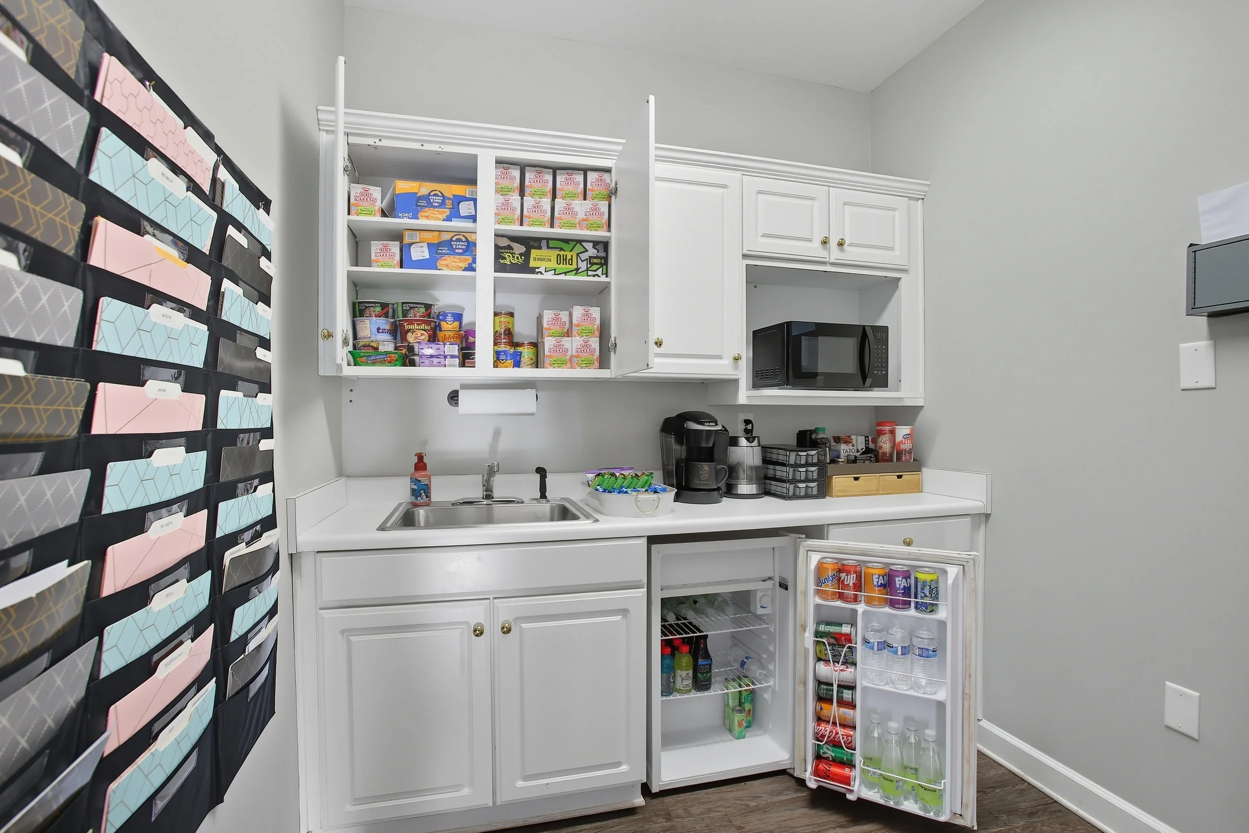 A small kitchenette with white cabinetry, an open pantry with food supplies, a microwave, a coffee maker, and a mini refrigerator filled with drinks.