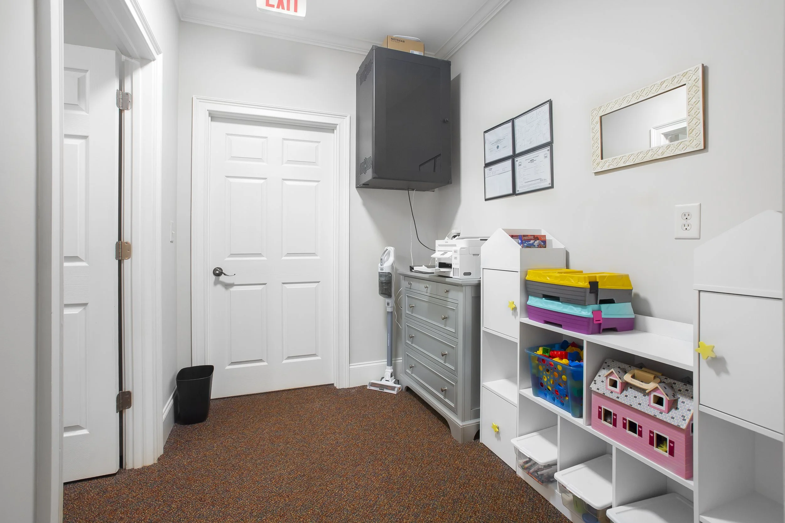 Office or classroom corner with a white storage unit, colorful toy boxes, a printer, a massage gun, picture frames on the wall, a mirror, and a door. Carpeted floor and a black waste bin.