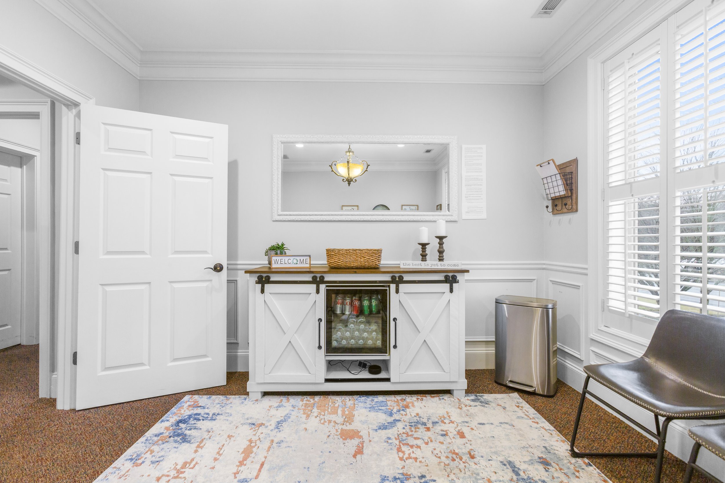 Bright living room corner with white walls and trim, a rustic white sideboard with sliding barn doors and glass-front storage, a large mirror framed in white above it, a small potted plant, a welcome sign, candles, and decorative items on top, two ch