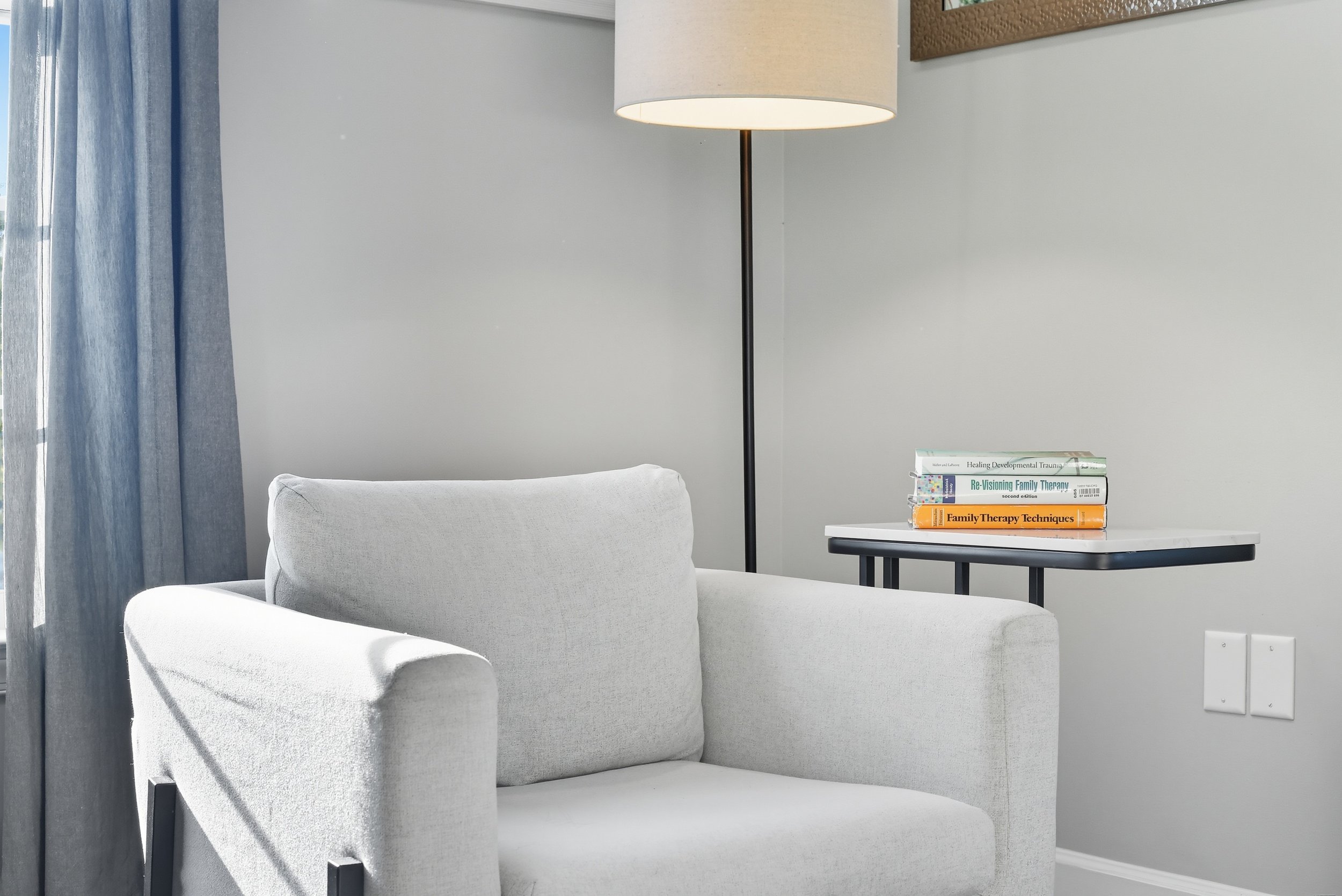 A white sofa next to a small table with books, a floor lamp, and a wall with a picture frame.