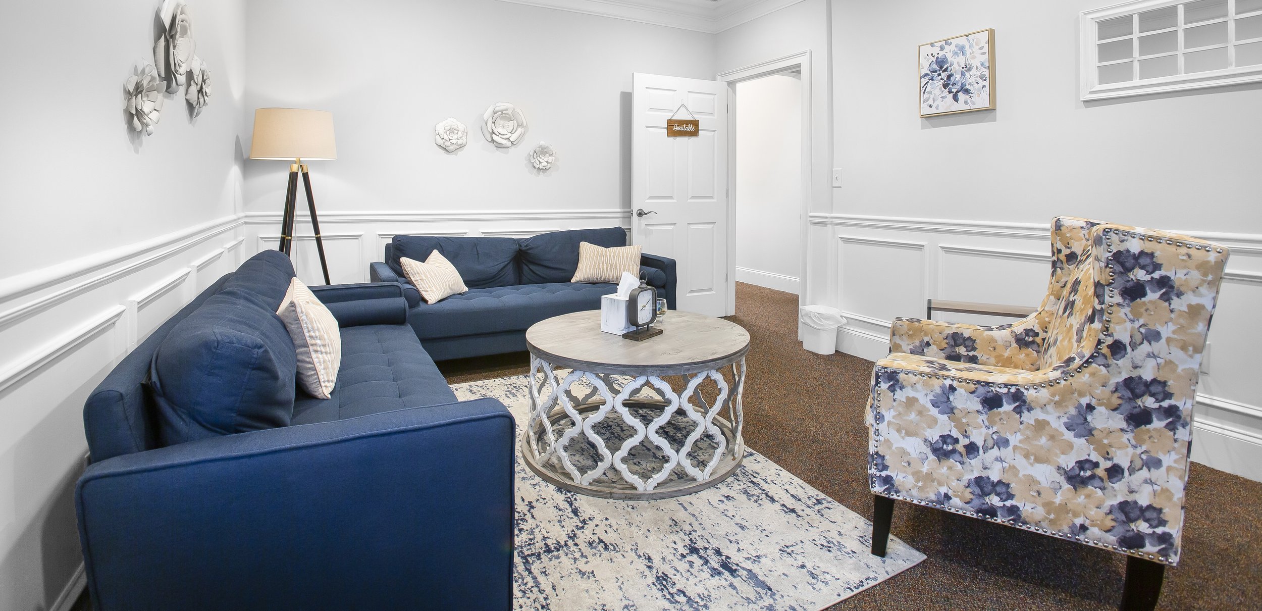 Living room with white walls, navy sofas, floral-patterned armchair, coffee table, and artwork on walls.