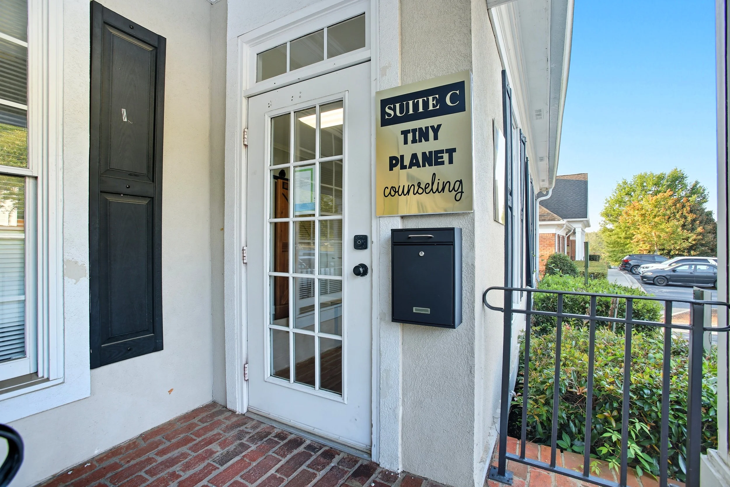 The entrance to Tiny Planet Counseling, Suite C, with a glass door, a black mailbox, and a sign on a white exterior wall, showing a parking lot with cars and trees in the background.