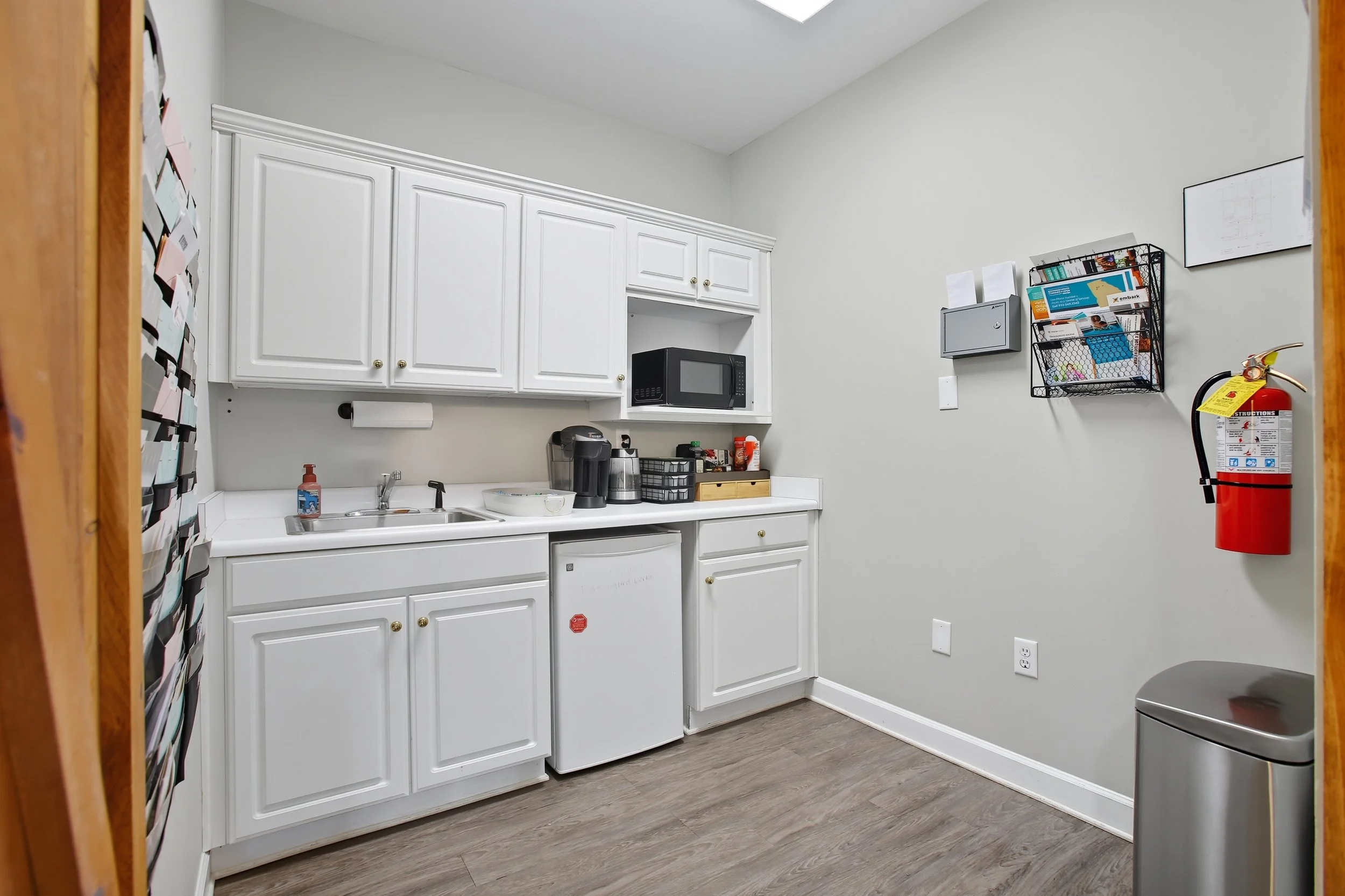 Small kitchenette with white cabinets, a microwave, and various appliances, including a coffee maker and mini refrigerator, against a light gray wall with wall-mounted storage and a fire extinguisher.