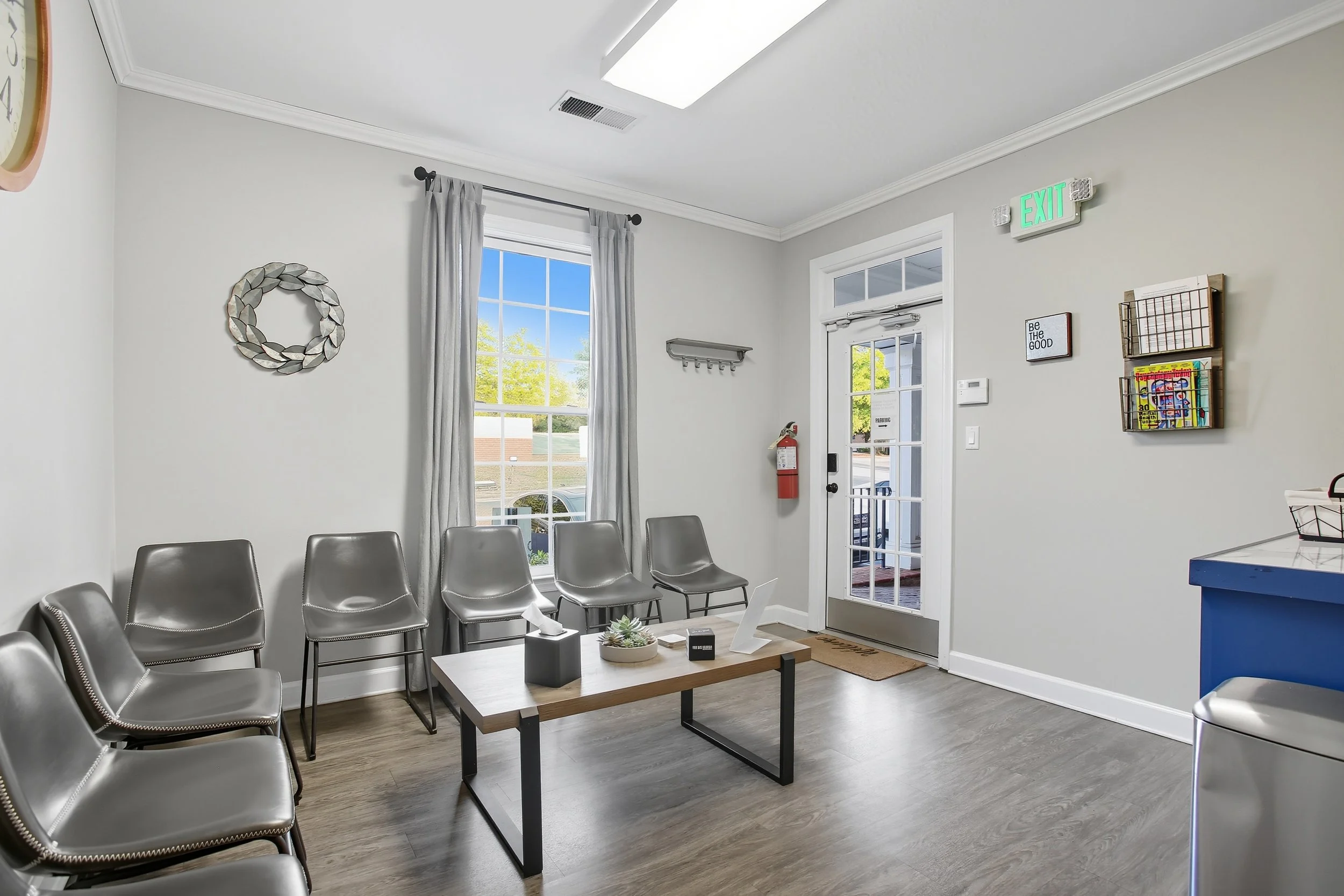 Waiting room with grey chairs, a wooden coffee table, and a window showing trees outside. The room has light grey walls, white crown molding, and a door with glass panes.