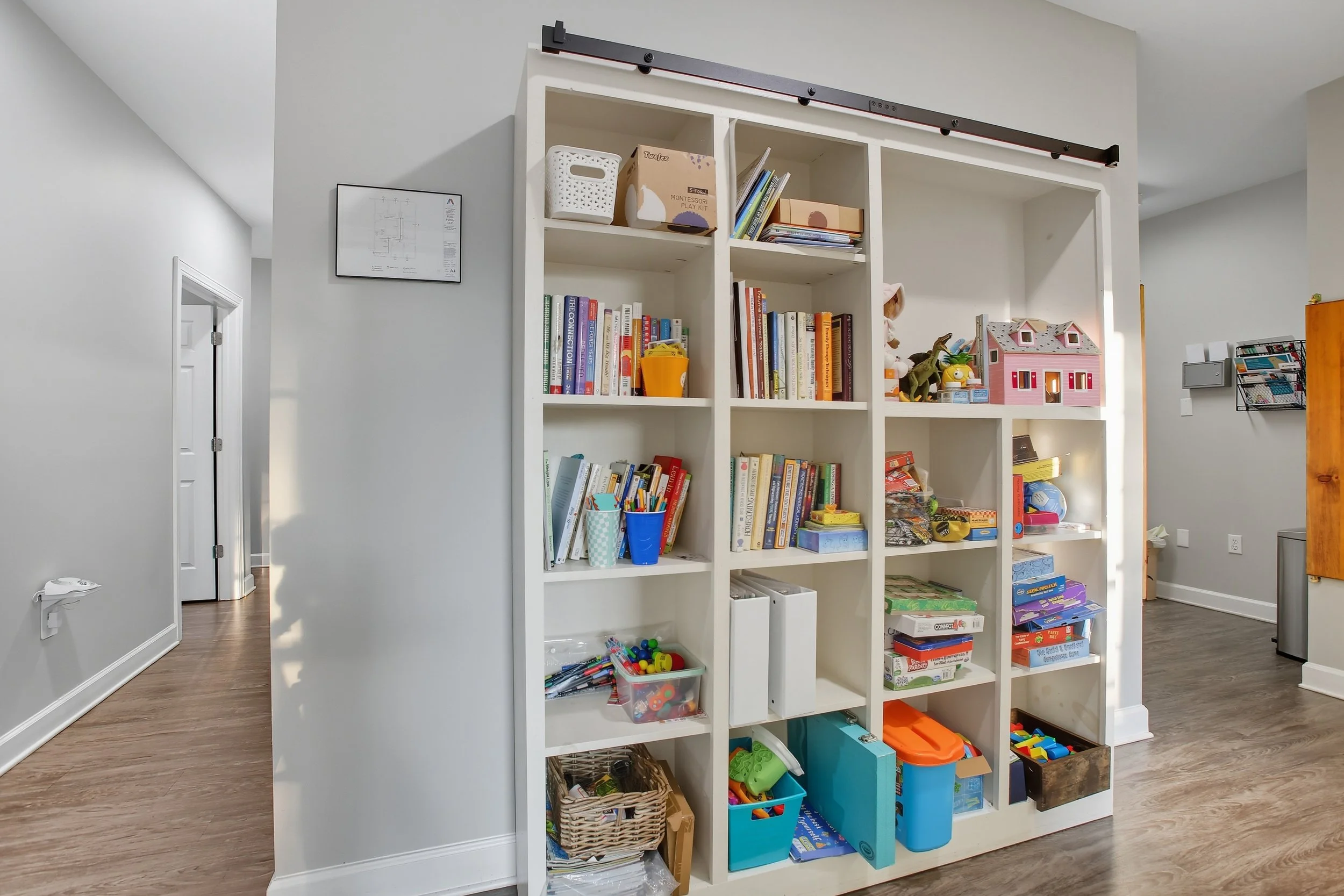 White bookshelf filled with children's books, toys, and games in a room with light gray walls and wooden floor.