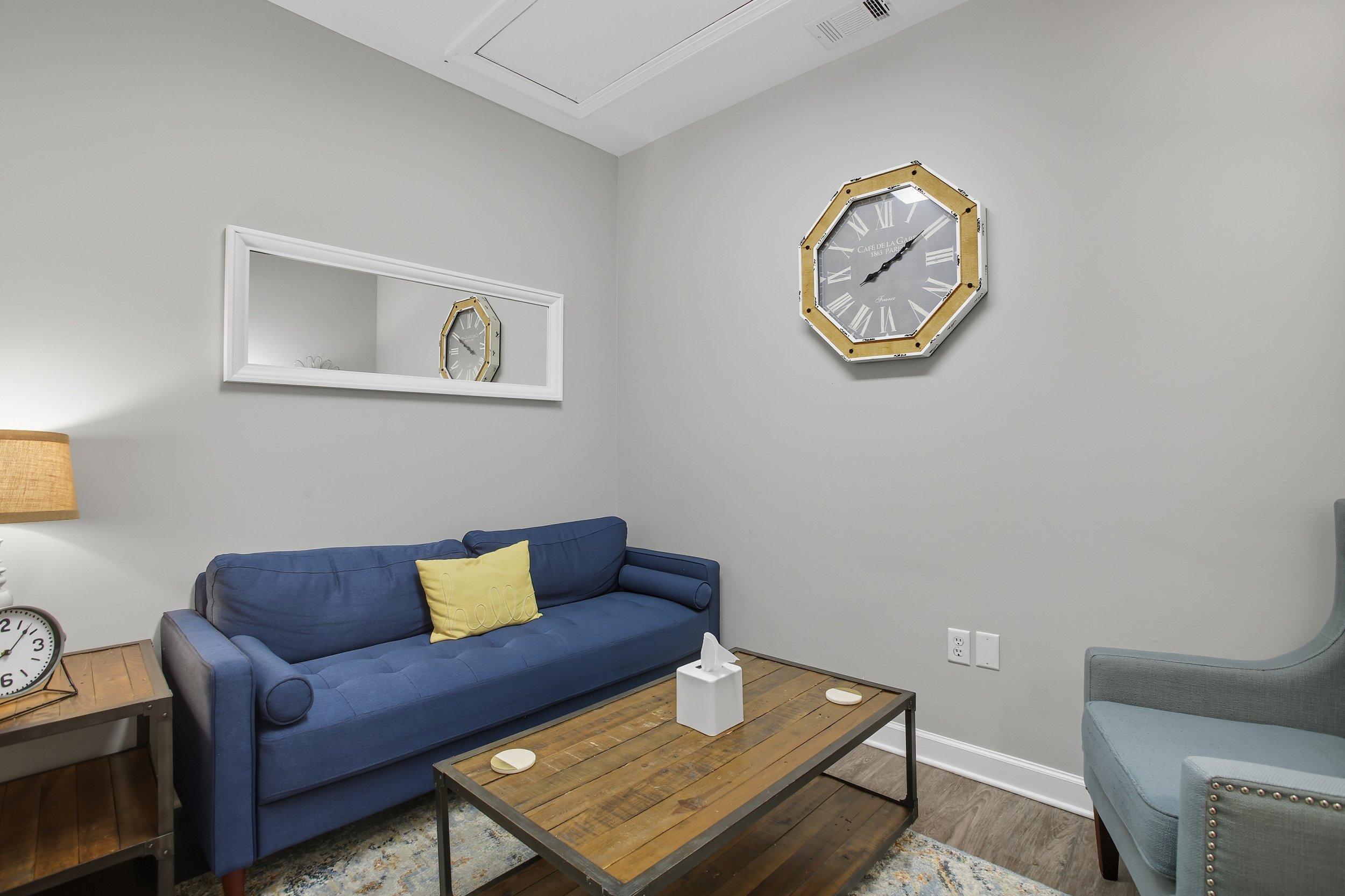 Living room with a blue sofa, a wall mirror, a wall clock, a wooden coffee table, a side table with a lamp and clock, a tissue box, and a remote in a modern decor style.