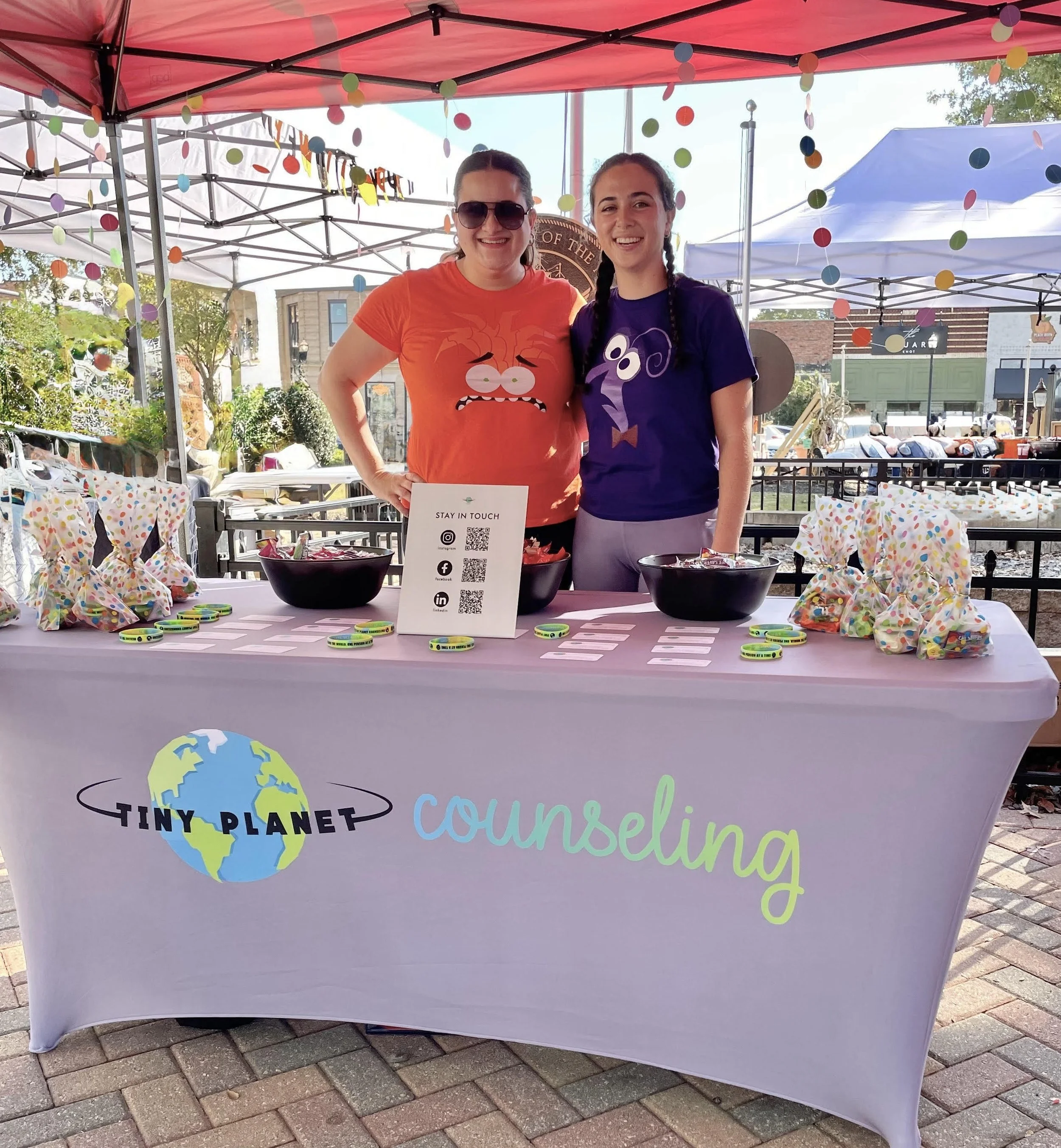 Two women standing behind a table with a sign for Tiny Planet Counseling at an outdoor event, decorated with colorful paper dots hanging from the tent, with bowls, small items, and a sign with QR codes on the table.
