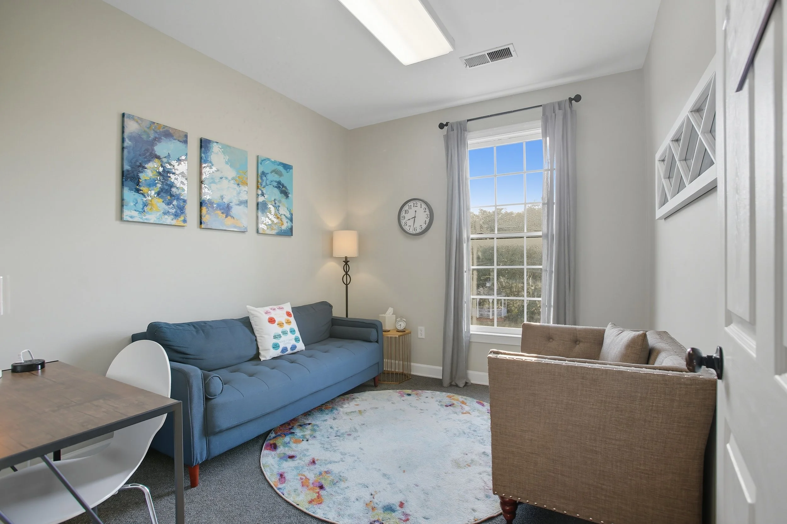 Living room with blue sofa, beige armchair, round colorful rug, abstract wall art, window with white curtains, wall clock, floor lamp, small side table, ceiling light, and partly open door.