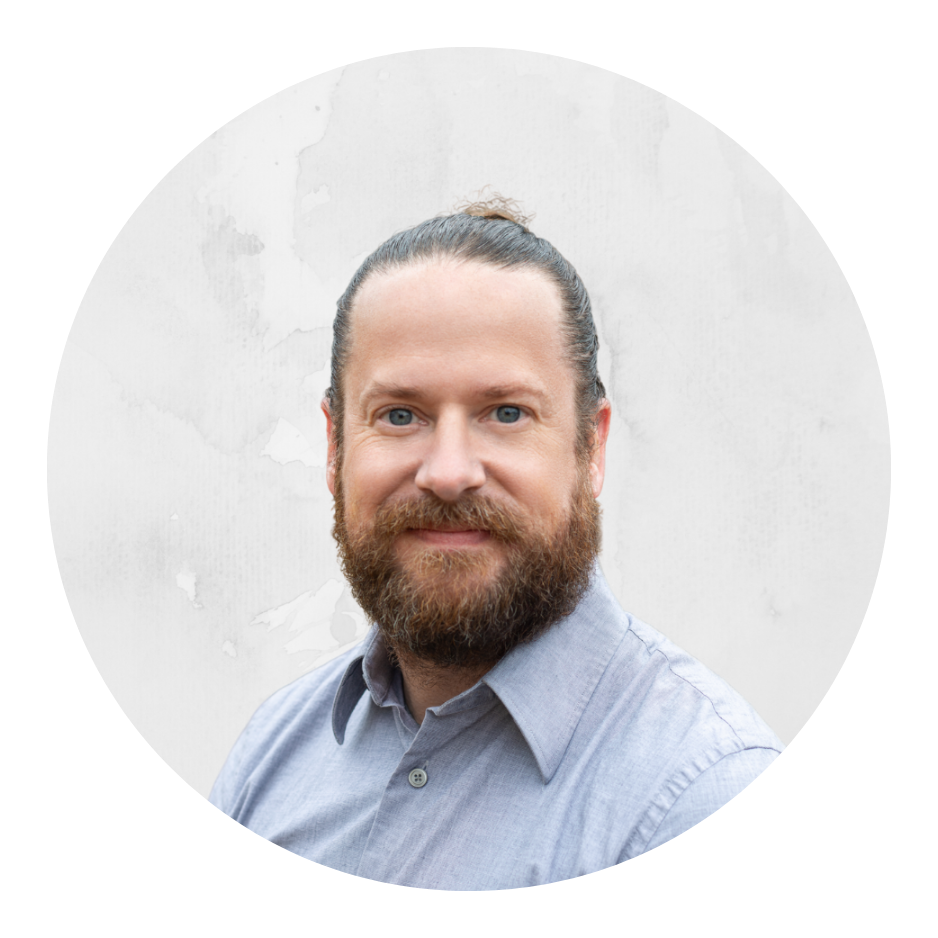 A man with a beard and tied-back hair, wearing a light gray button-up shirt, standing outdoors with trees in the background.