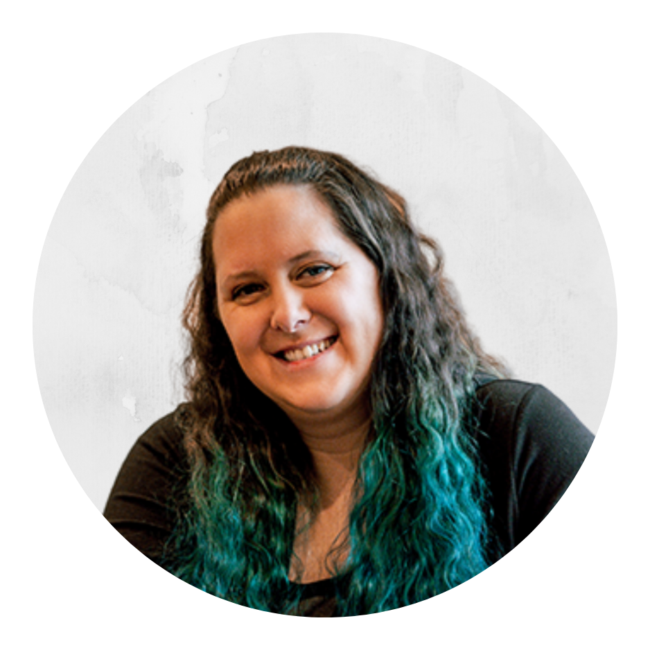 Woman with long wavy hair that is dark at the top and transitions to teal at the ends, smiling while sitting indoors.