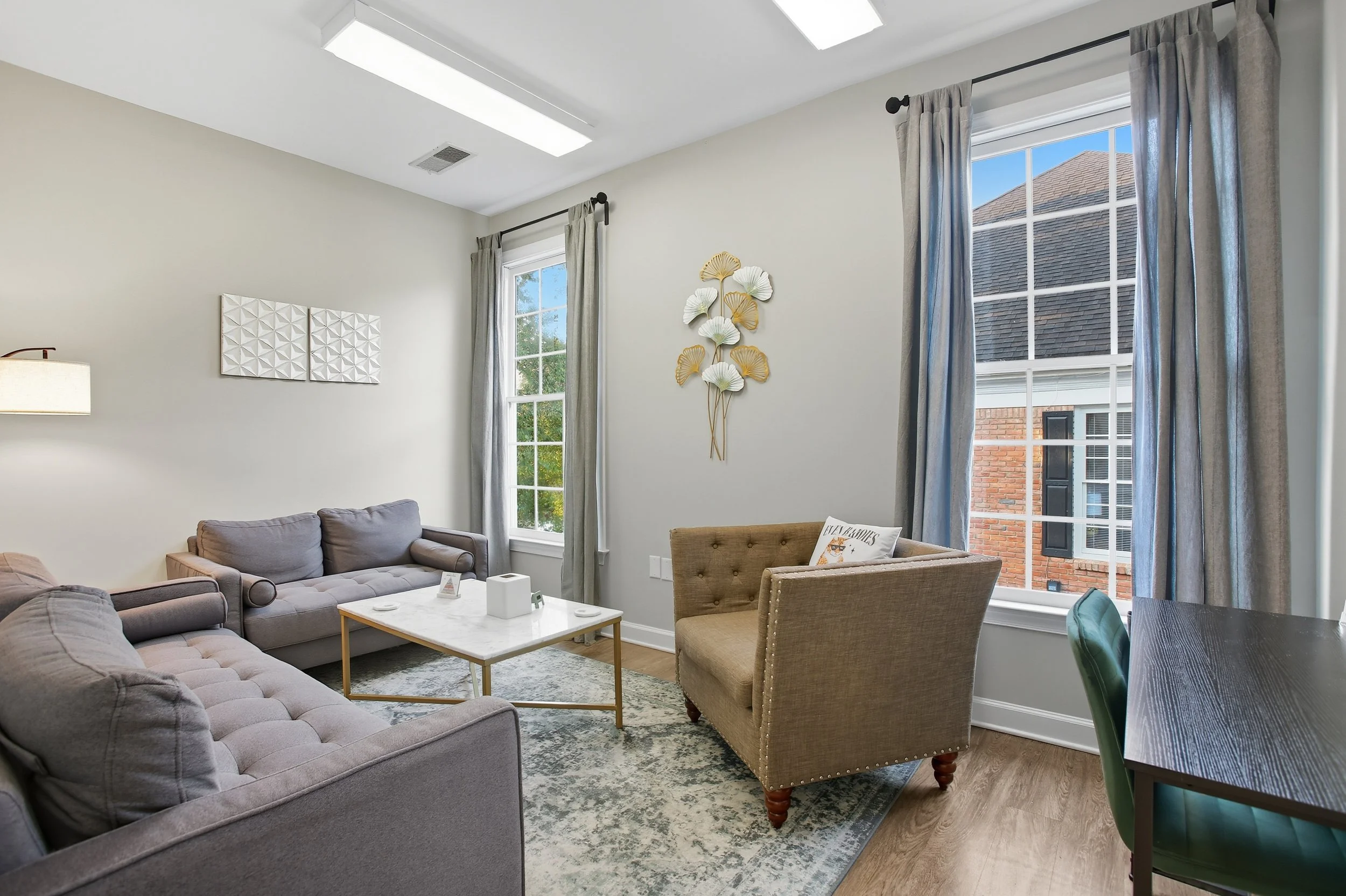 Living room with gray sofas, a beige armchair, a white marble coffee table, large windows with curtains, and decorative wall art.