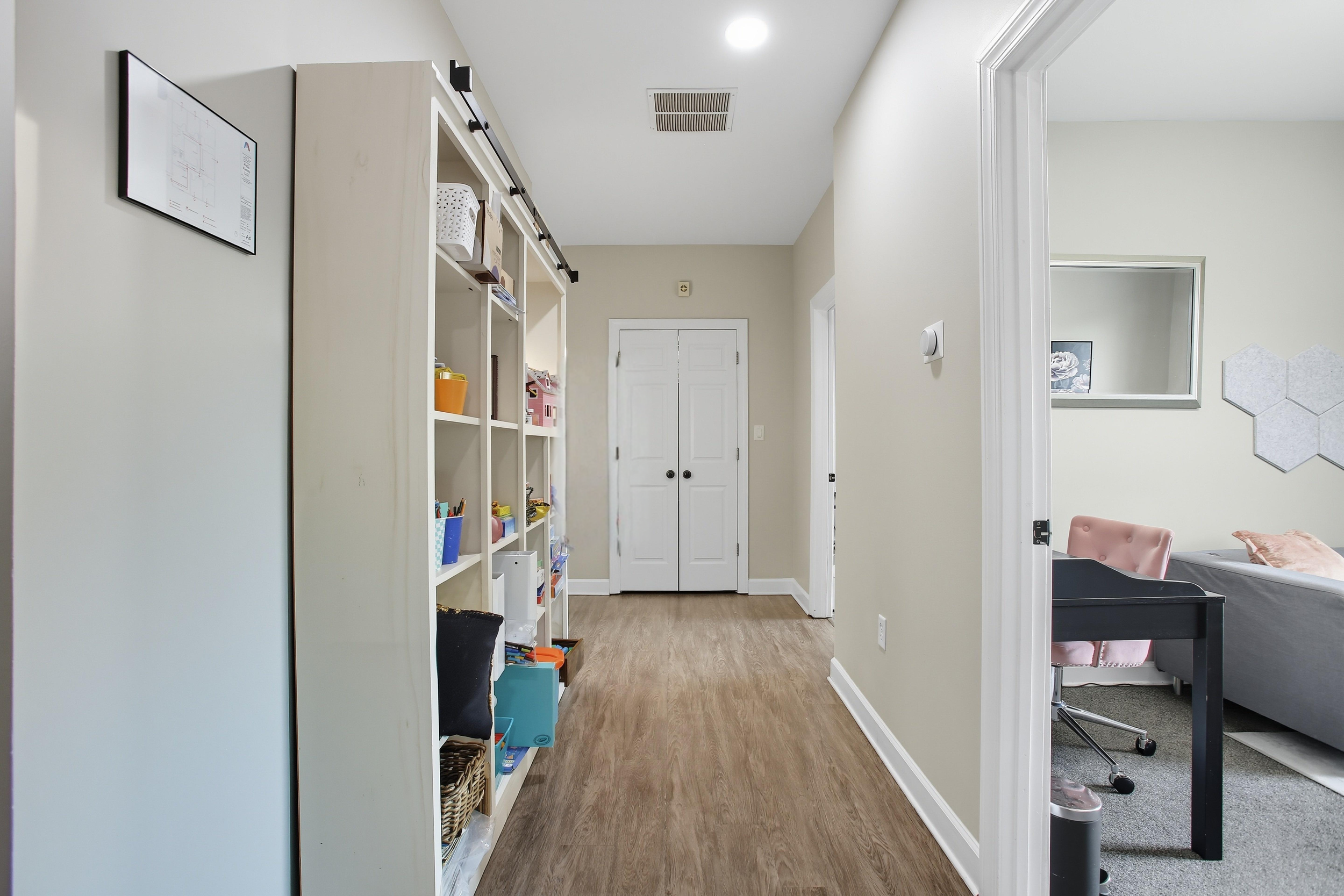 Hallway with bookshelf on the left and an open doorway on the right leading to a room with a pink chair and picture on the wall.