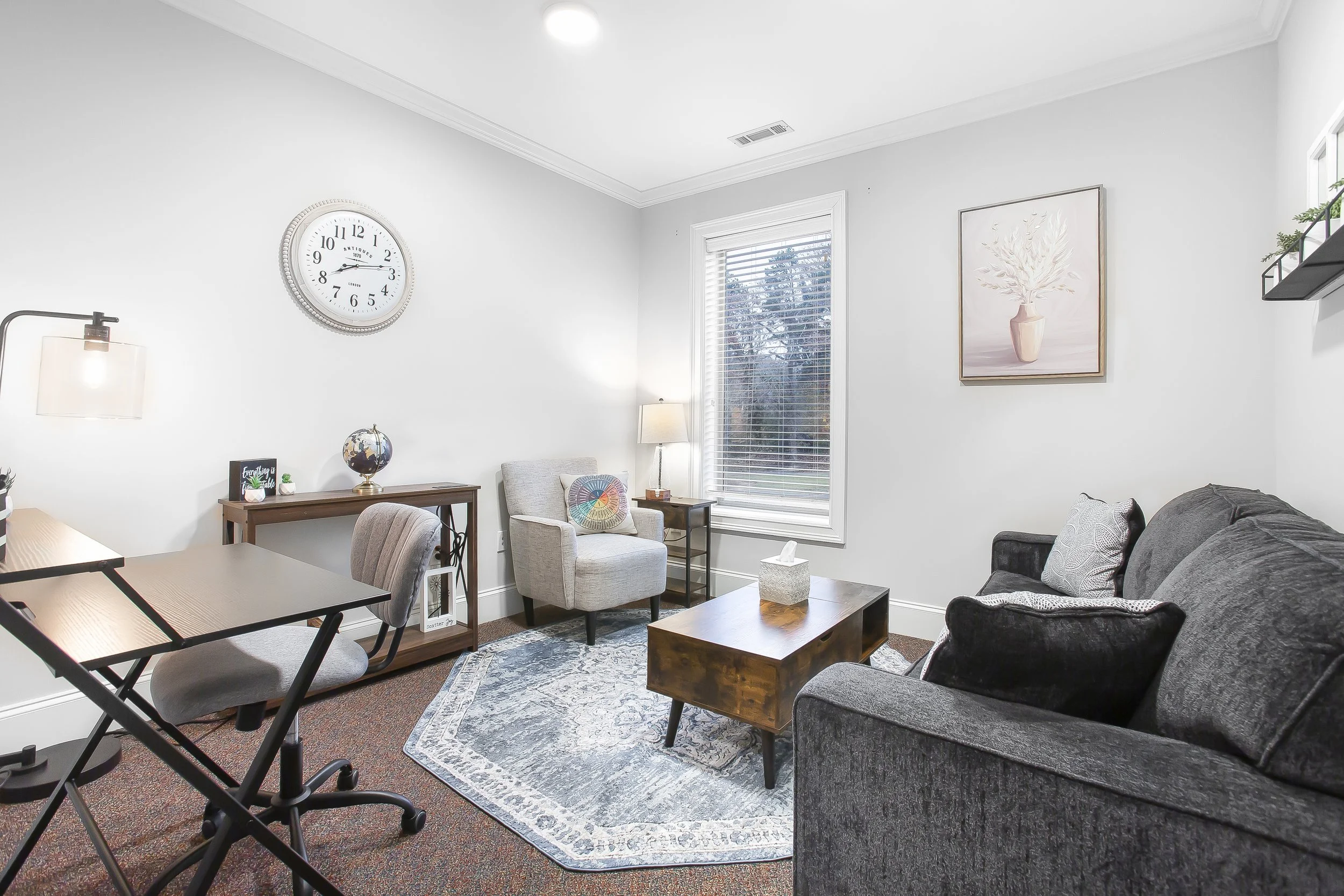 Living room with a gray sofa, beige armchair, wooden coffee table, patterned rug, window with blinds, scholarly wall art, clock, side table with lamp, and wall shelf with plants.