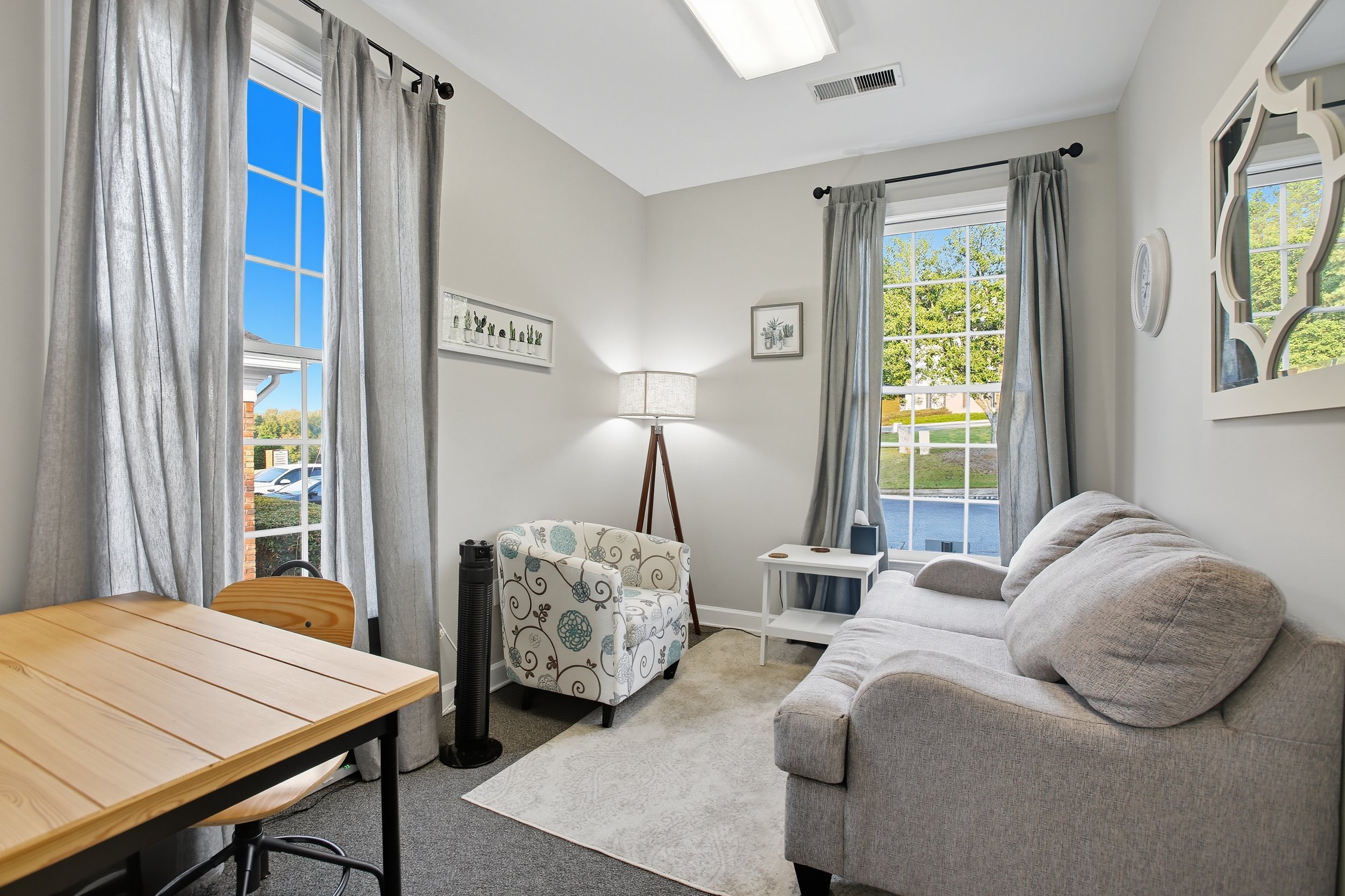 A living room with two large windows with gray curtains, a white sofa, a small white side table, a patterned armchair, a floor lamp, and a wooden dining table with a chair. The room has white walls and carpeted floor, with picture frames and a mirror