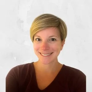 Portrait of a woman with short blonde hair smiling, wearing a dark top, against a light background.