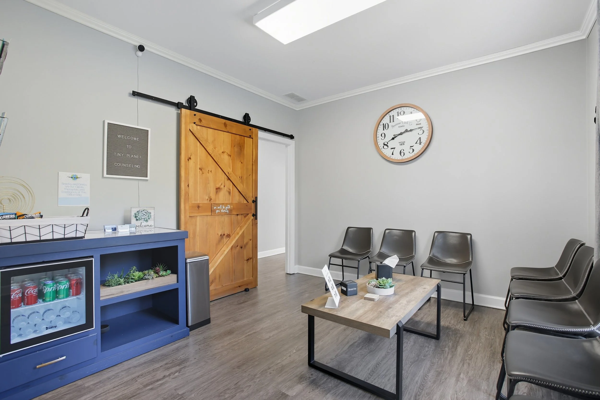 Waiting room with four black chairs along the wall, a wooden coffee table with a potted succulent and tissue box, a blue cabinet with drinks, a wooden sliding door, and a clock on the wall showing 2:18.
