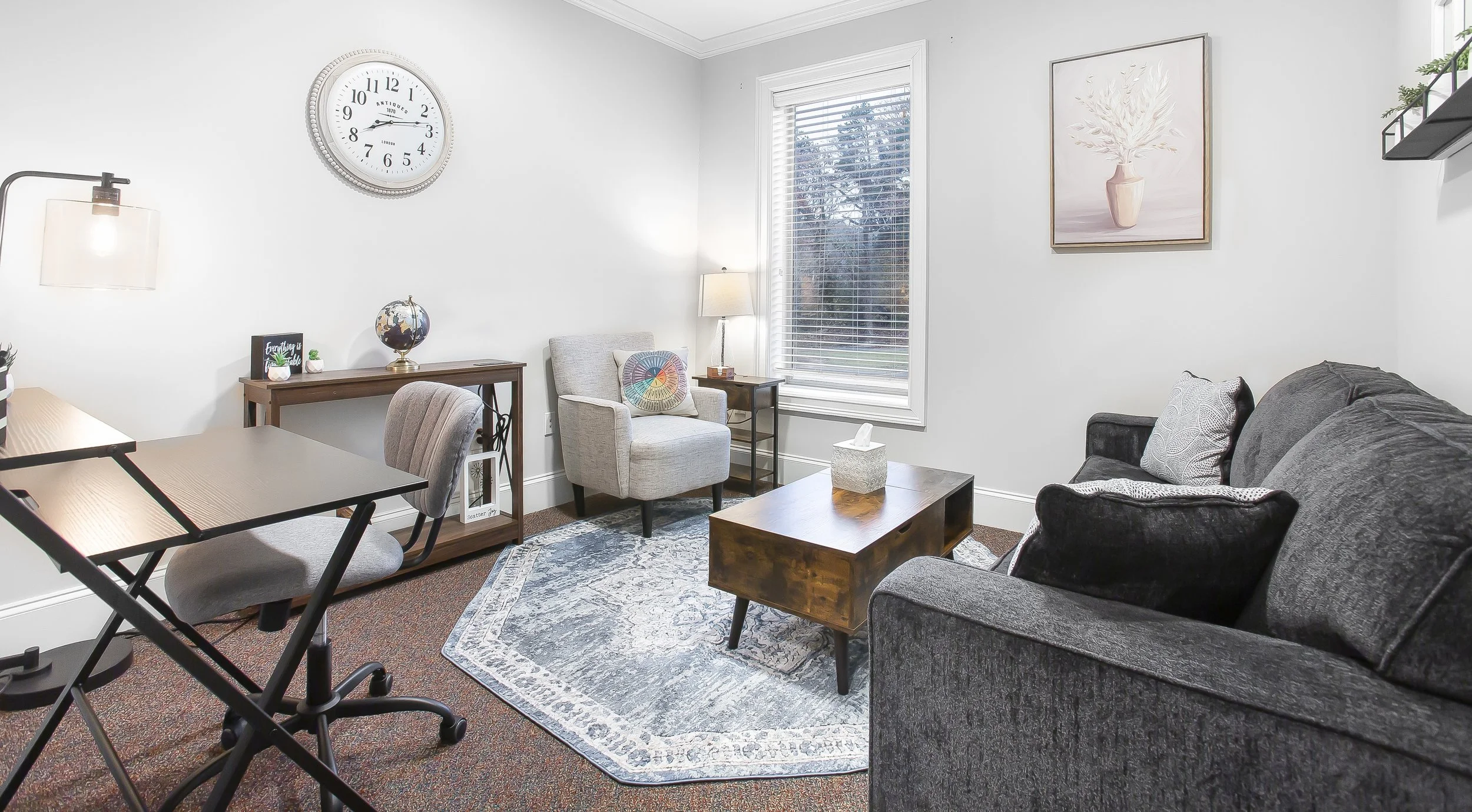 Living room with gray sofa, armchair, wooden coffee table, area rug, window with blinds, wall clock, framed artwork, table lamp, floor lamp, and decorative items.