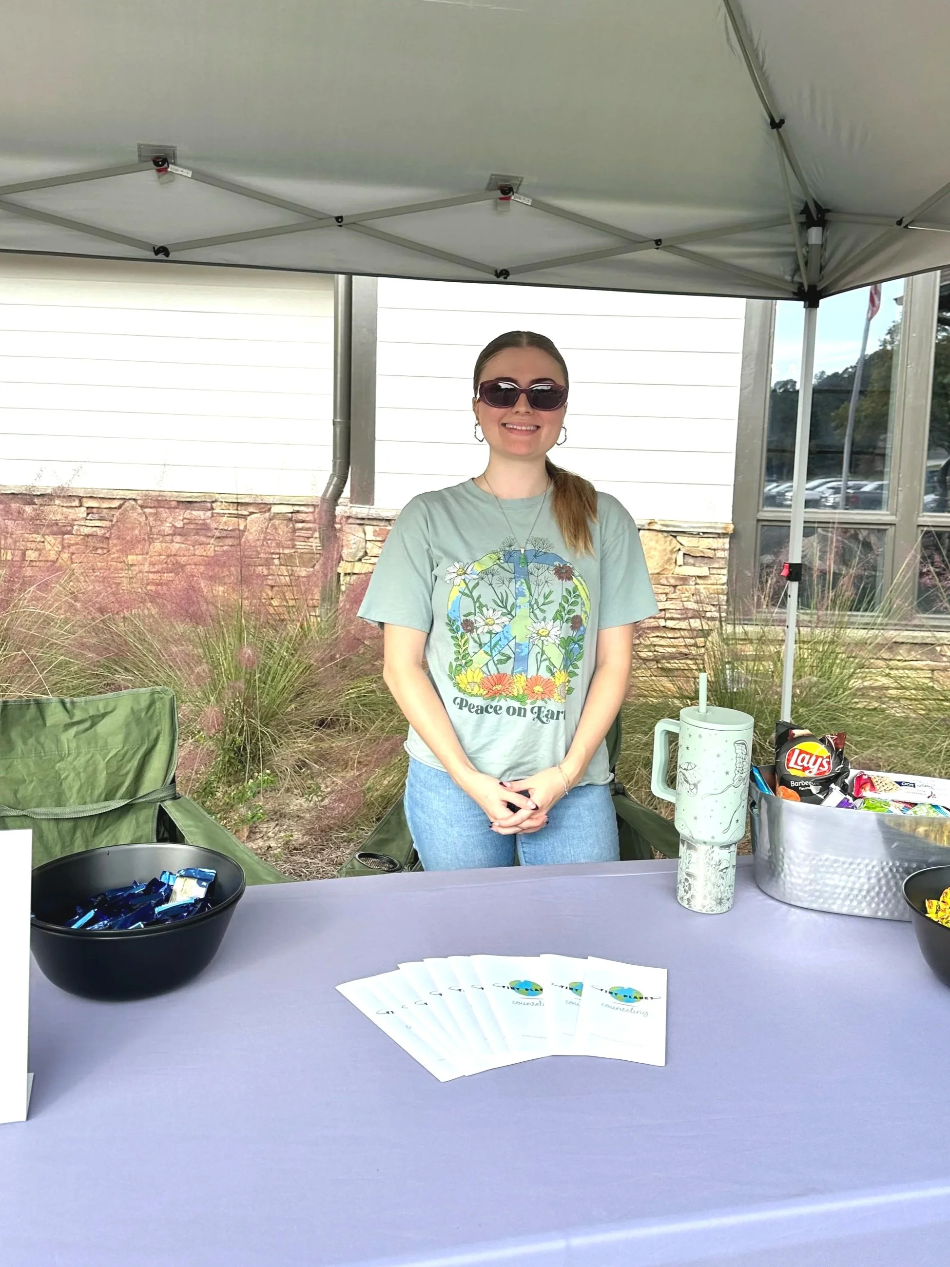 Woman with sunglasses smiling standing behind a table with snacks and pamphlets, under a canopy outdoors.