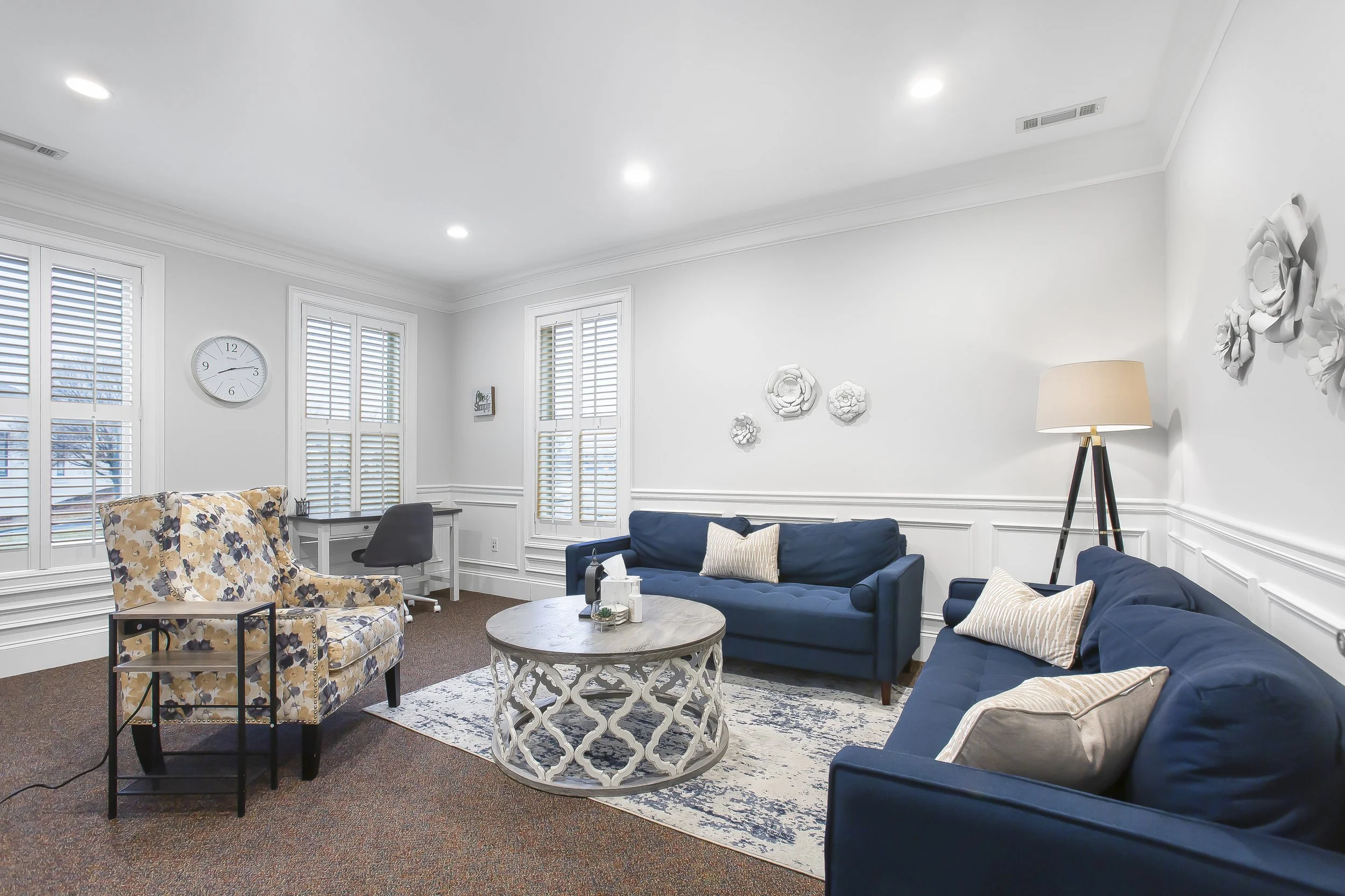 Living room with two blue sofas, a floral armchair, a round coffee table, a floor lamp, a desk with a chair, and white wall decor, windows with shutters, and a clock on the wall.