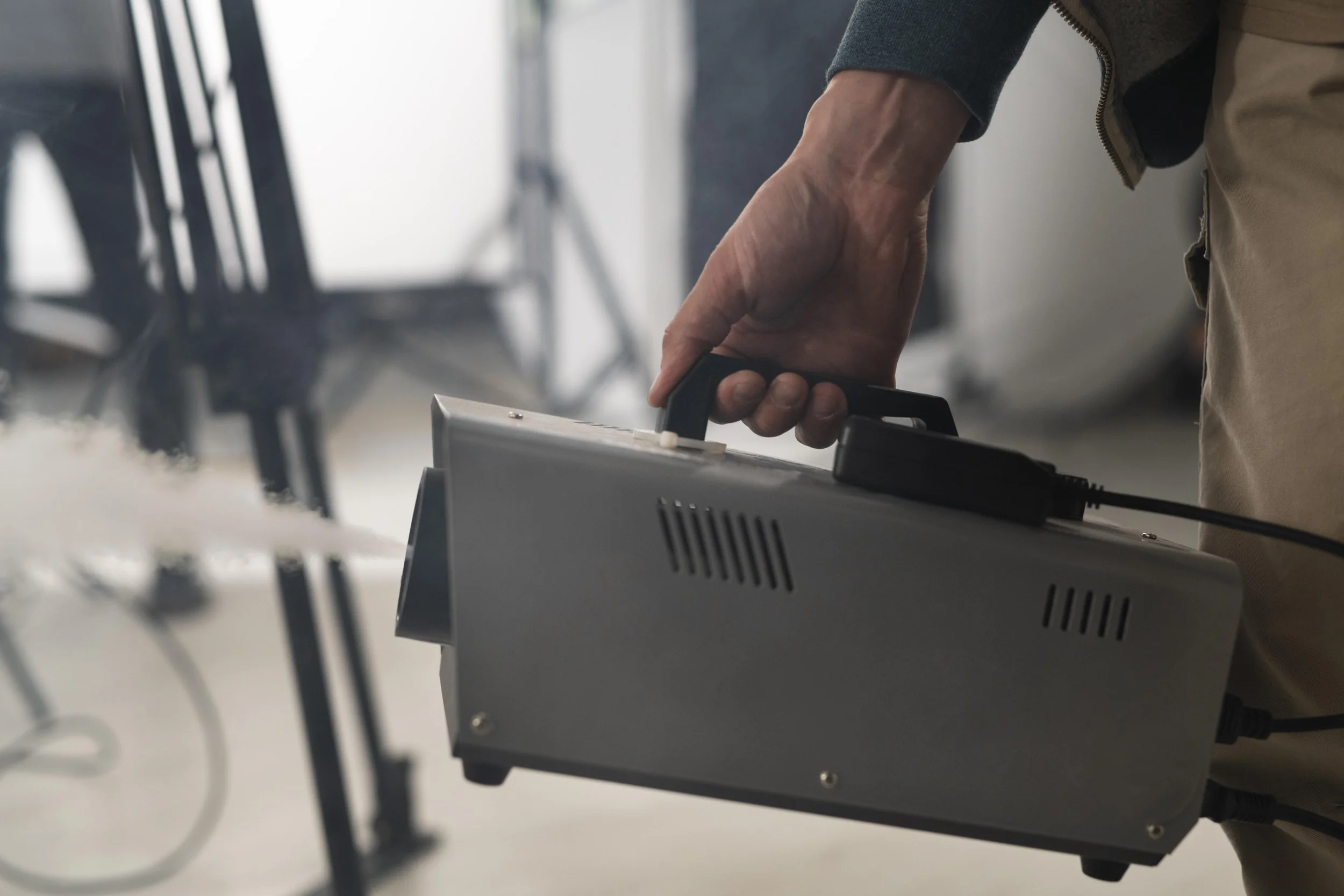 A person holding a gray device with a handle, possibly a fogging or spraying machine, with a cord attached.