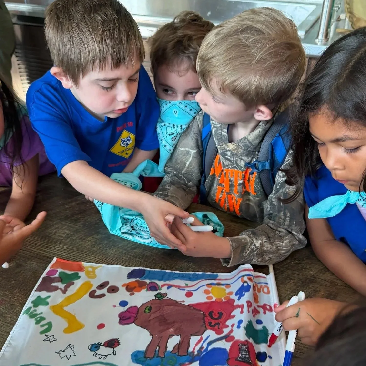 Group of children gathered around a table looking at a colorful drawing of a horse on a white cloth, with various figures and letters, some children holding markers and one child wearing a camouflage jacket and a blue bandana.