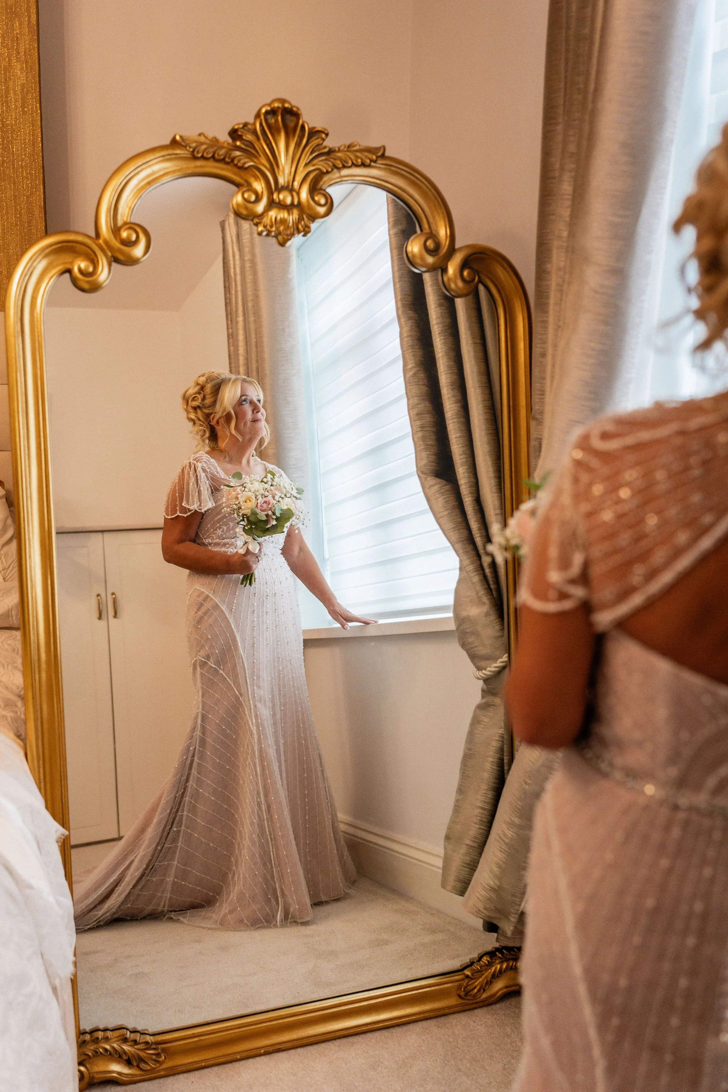 A woman in a long, elegant gown holding a bouquet of flowers, looking at her reflection in a large, ornate gold mirror.