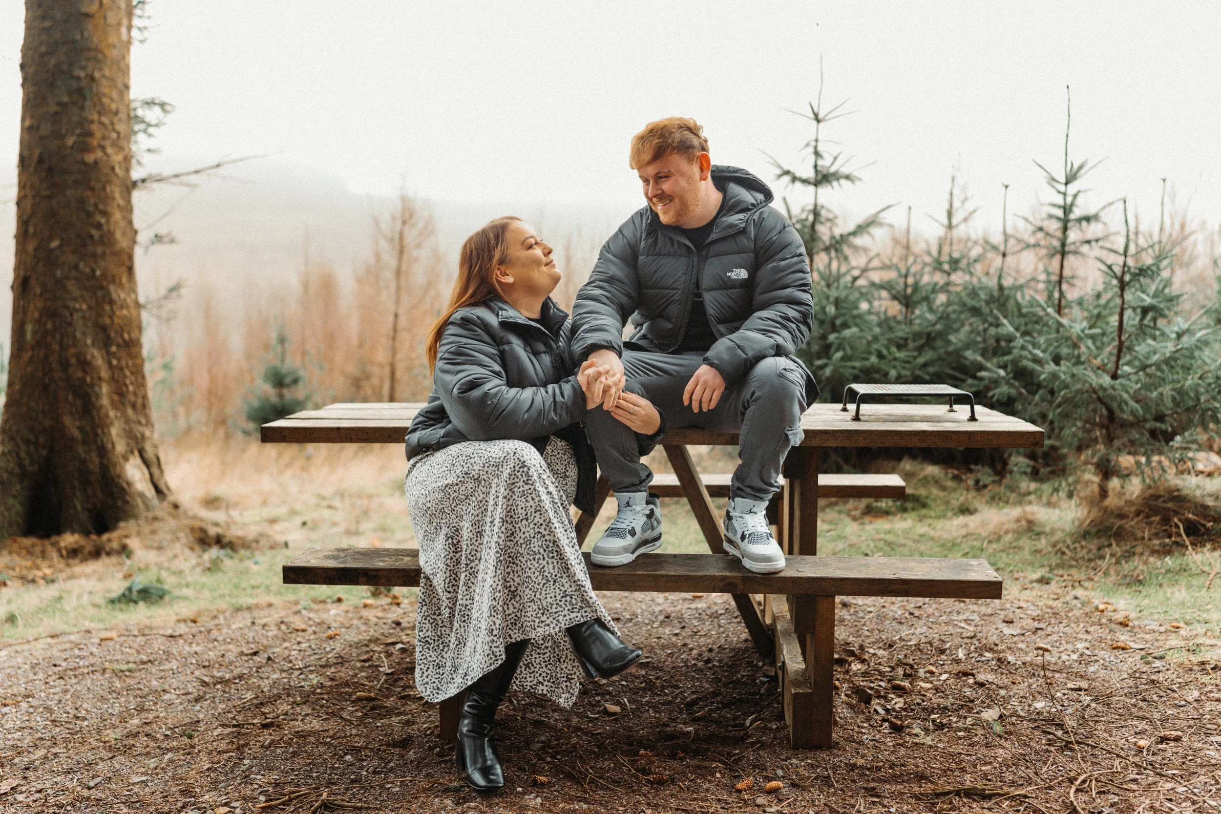 A couple sitting on a picnic table outdoors in a forest, holding hands and looking at each other smiling.
