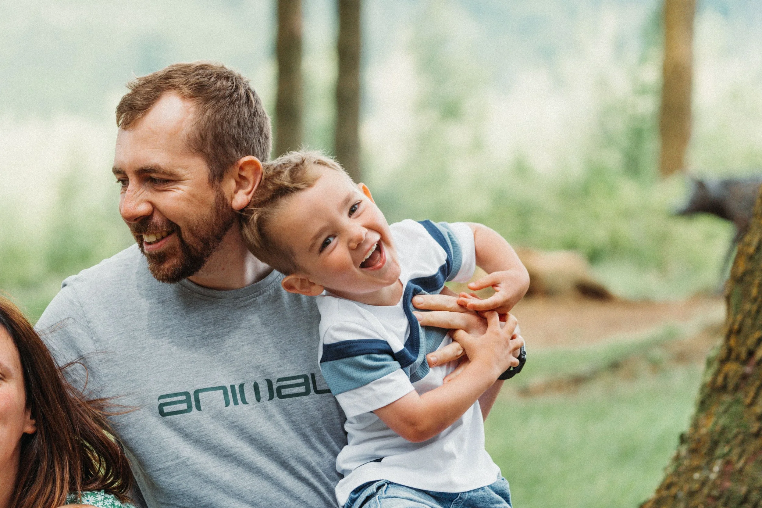 A man holding and playing with a smiling young boy outdoors in a wooded area.