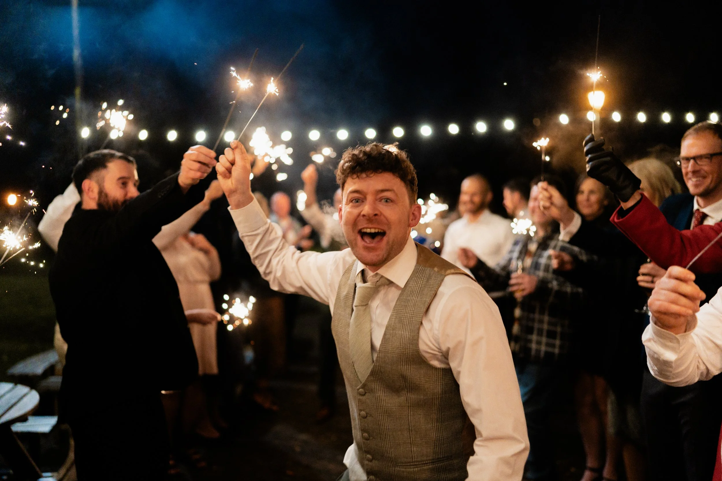 Celebration of New Year's Eve with people holding sparklers and smiling, under string lights at night.