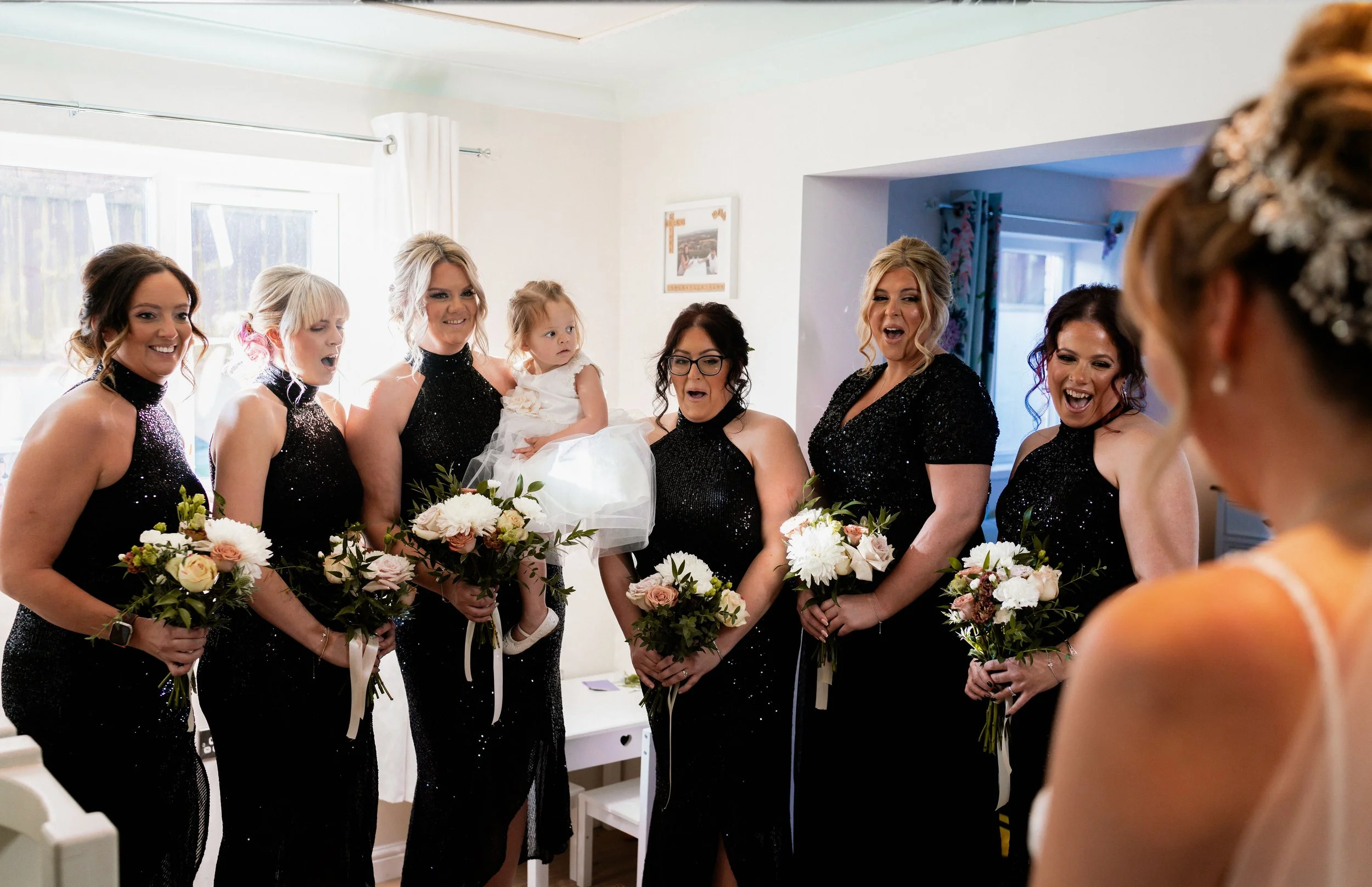 A bride with her back to the camera in a white dress, facing her bridesmaids who are holding bouquets and reacting with surprise and joy in an indoor setting.