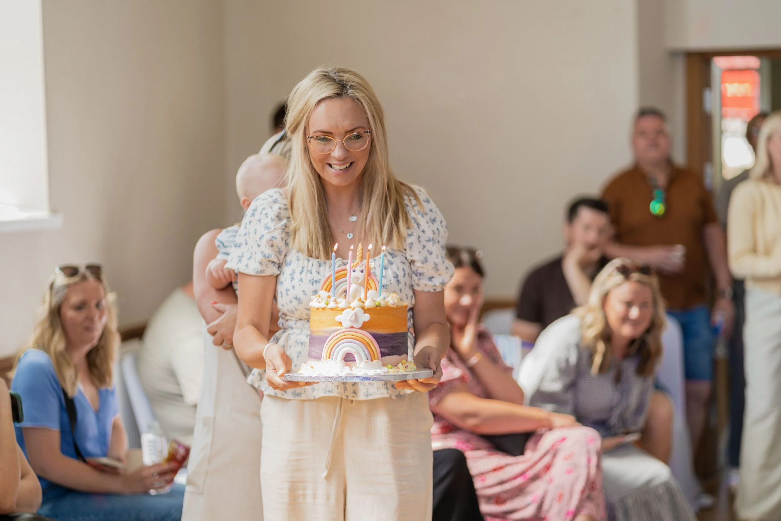 Woman holding a colorful unicorn-themed birthday cake with lit candles, smiling at a celebration gathering.