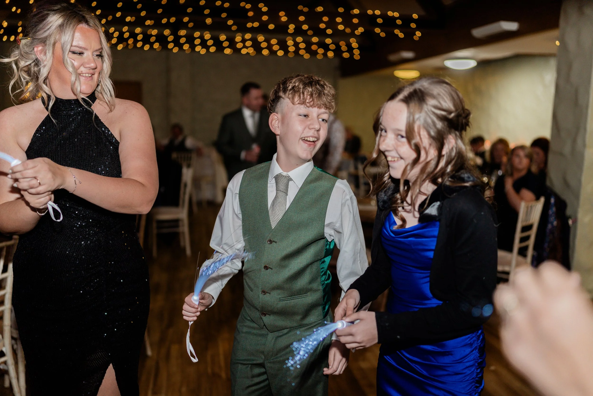 Three people, two girls and one boy, smiling and holding sparklers at an indoor celebration or party, with other people seated in the background.